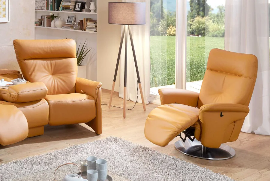 Wohnzimmer mit zwei orangefarbenen Ledersesseln, davon einer mit Fußstütze ausgeklappt, auf Teppich vor Fensterfront.
