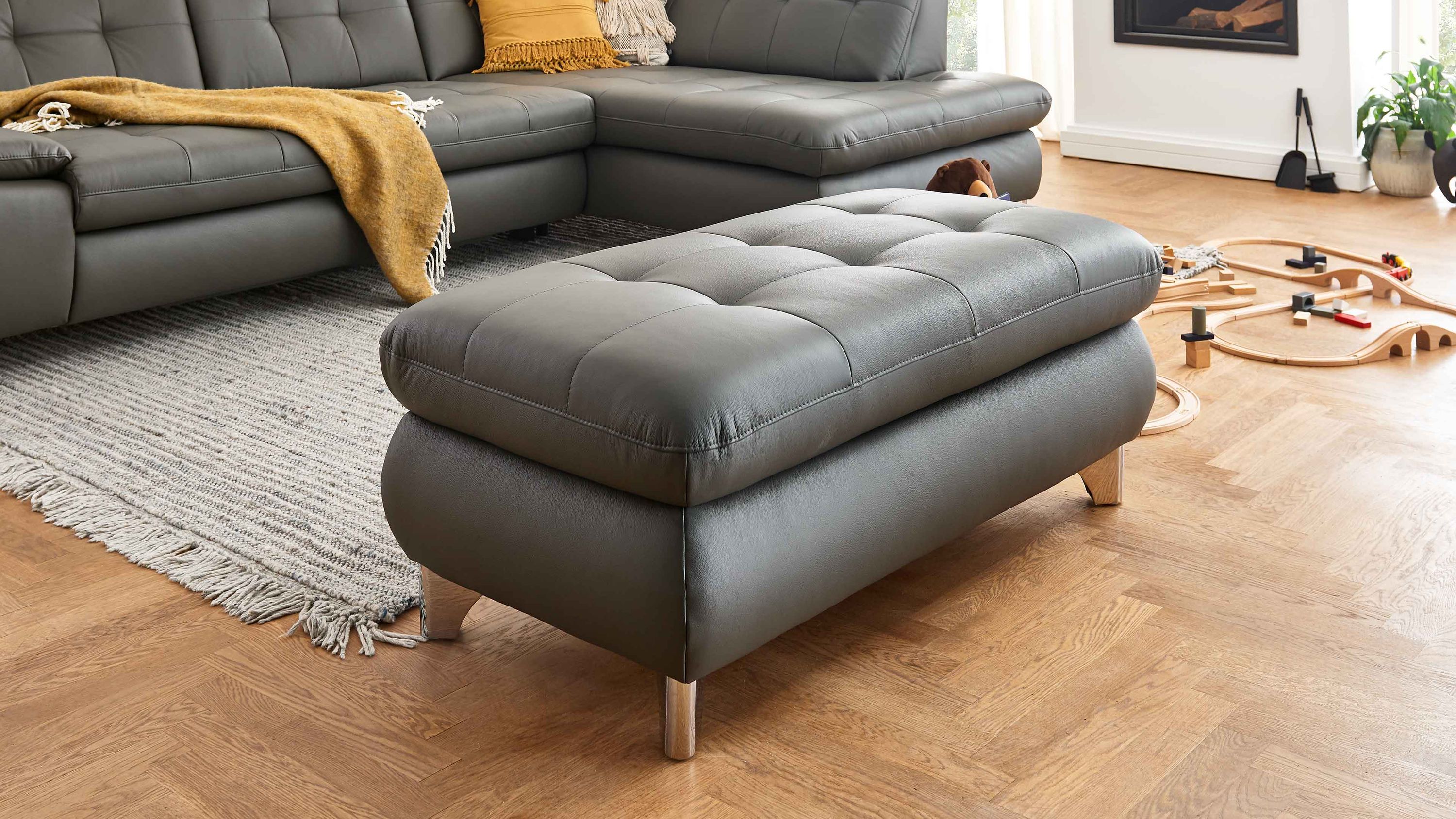 Rechteckiger grauer Hocker mit gepolstertem Deckel und glänzenden Winkelfüßen vor grauem Sofa