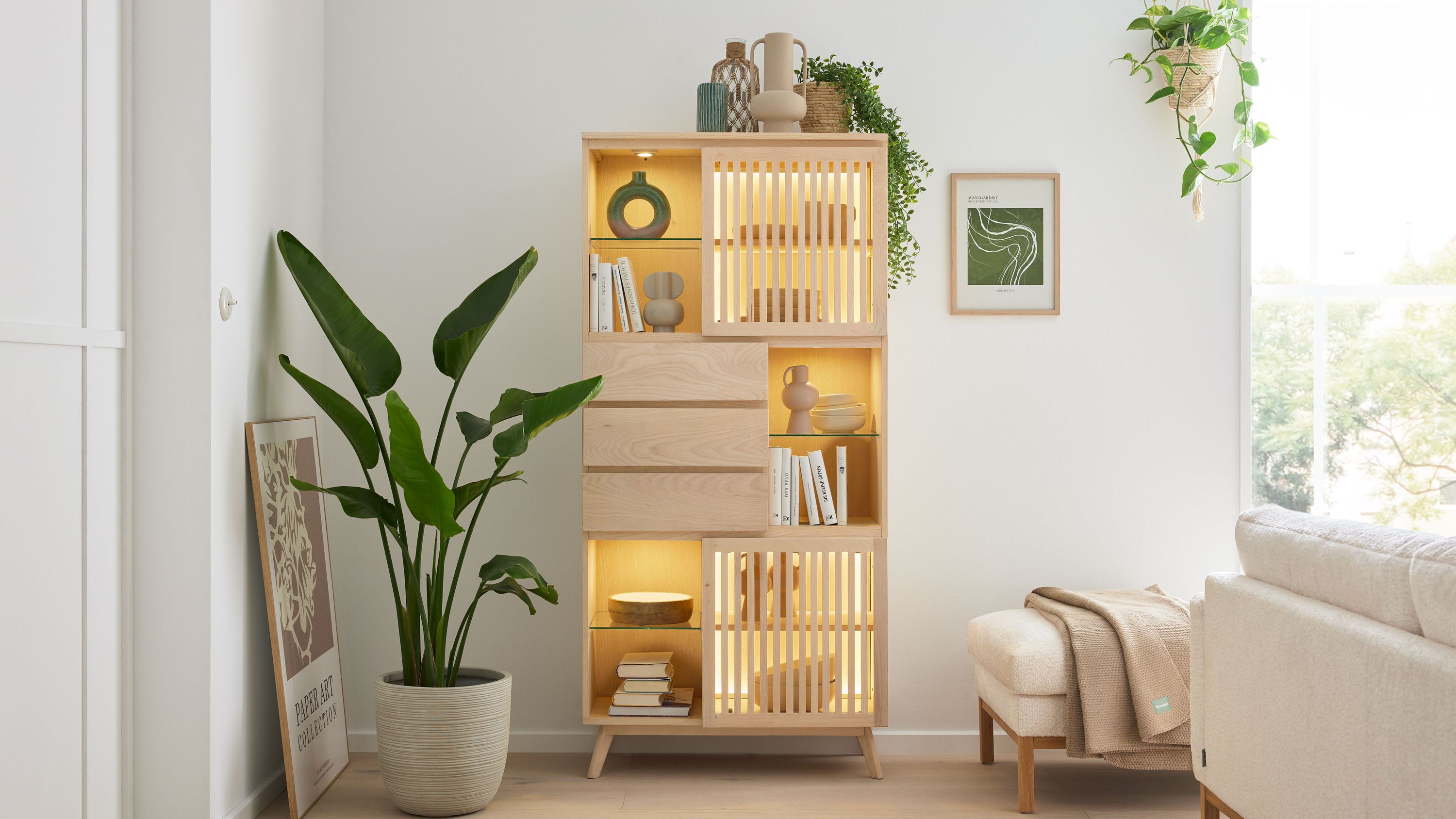 Vitrine mit Türen und Schubladen in hellem Holz Helle Vitrine mit zwei Türen und drei Schubladen beleuchtet neben Pflanze und Sofa