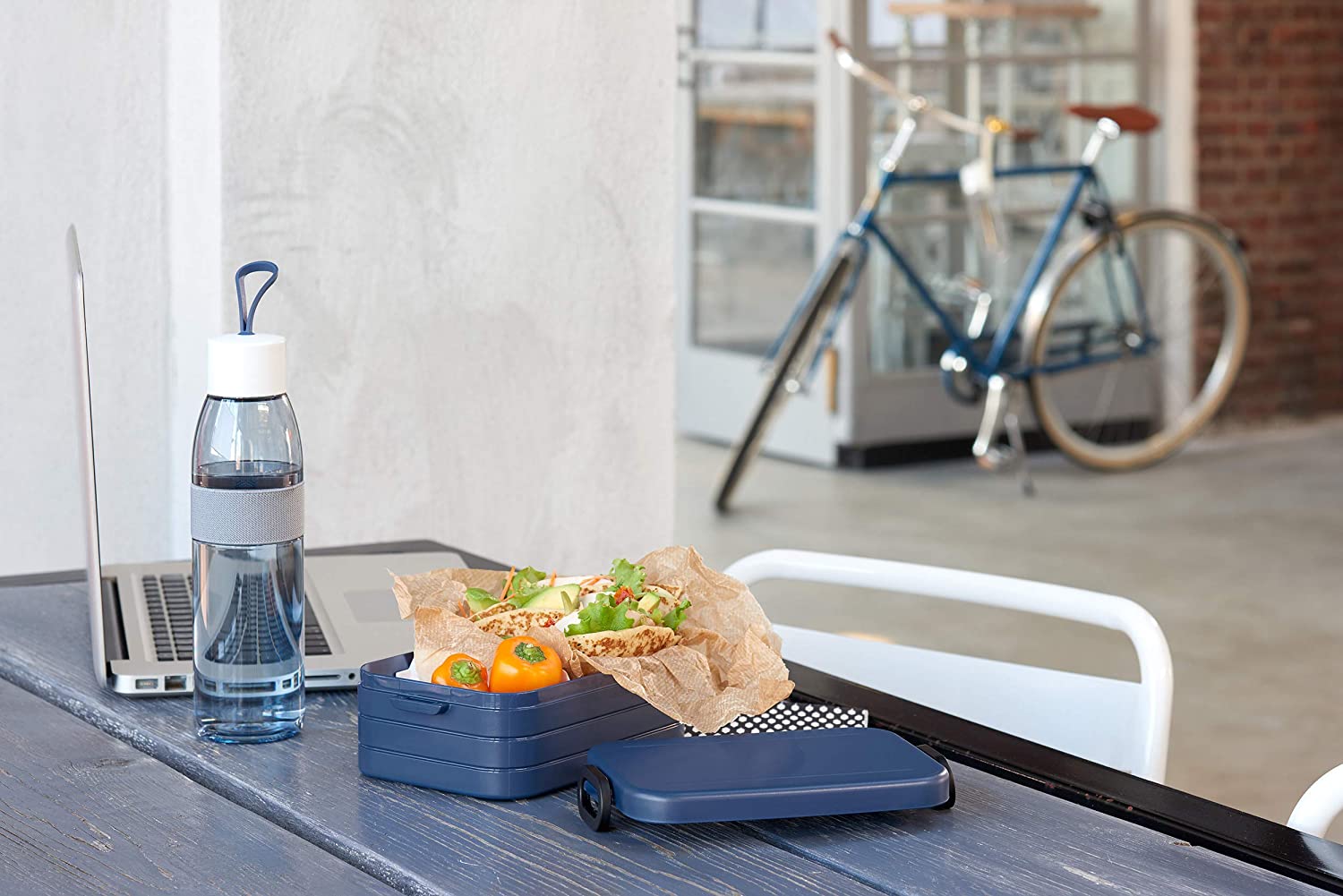 Blaues Mepal-Lunchbox-Set mit Sandwich und Trinkflasche auf Tisch, Laptop und Fahrrad im Hintergrund