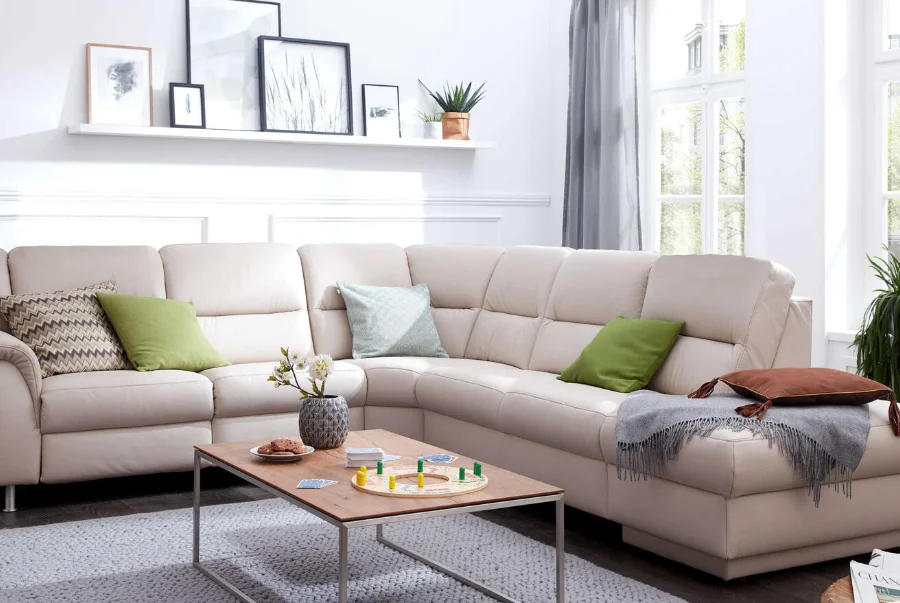 Helles Wohnzimmer mit beiger Eckcouch, grünen Kissen, Holztisch mit Gesellschaftsspiel und dekorativen Wandbildern.