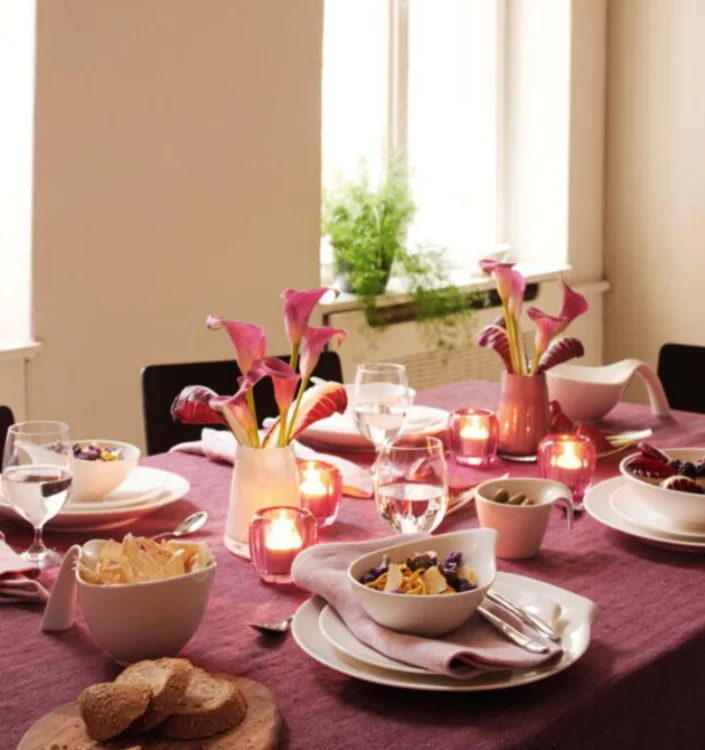 Festlich gedeckter Esstisch mit lila Tischdecke, Kerzen, Blumenvasen, Geschirr und Gläsern in gemütlicher Atmosphäre.