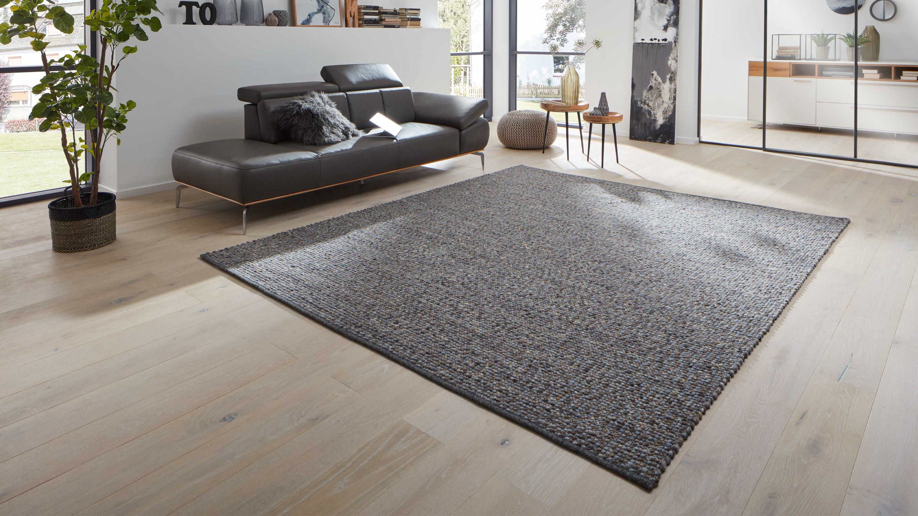 Großflächiger grauer Teppich mit grober Struktur in modernem Wohnzimmer mit schwarzem Ledersofa und hellen Holzboden