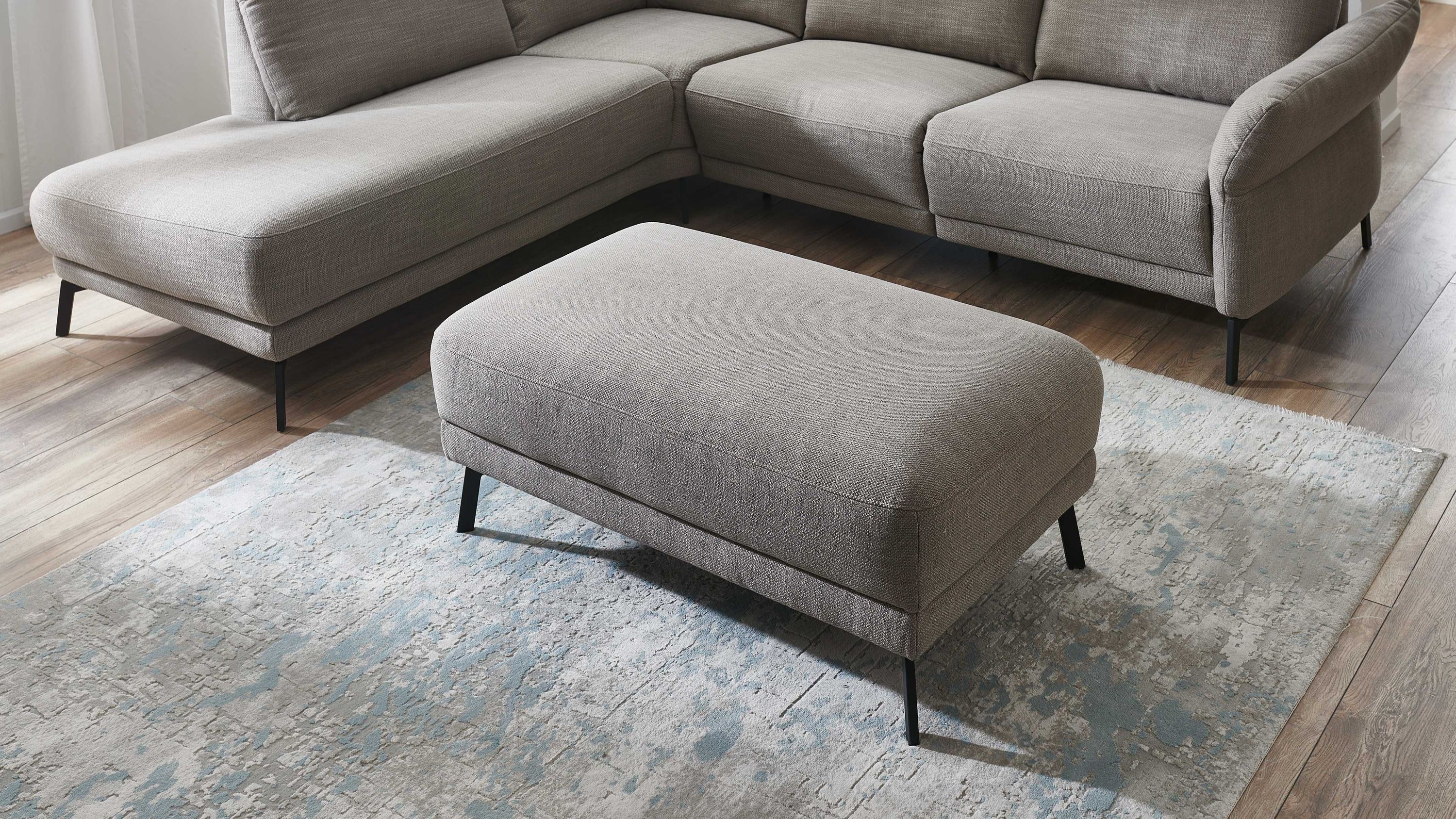 Rechteckiger Polsterhocker mit taupefarbenem Stoff und schwarzen Metallfüßen vor grauem Ecksofa