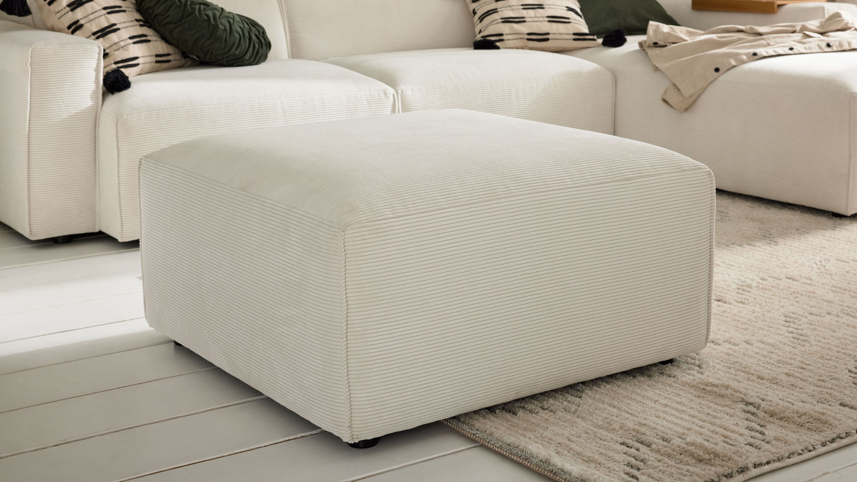 Weißer quadratischer Polsterhocker mit Cordbezug Quadratischer Polsterhocker in weiß mit feiner Cordstruktur in einem Wohnzimmer vor einem Sofa