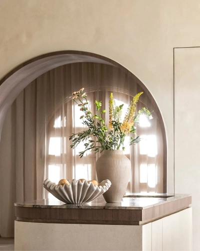 Dekorative Blumenvase und Obstschale auf eleganter Kücheninsel. Große Vase mit frischen Blumen und Obstschale auf Kücheninsel vor rundem Fenster mit lichtdurchlässigem Vorhang.