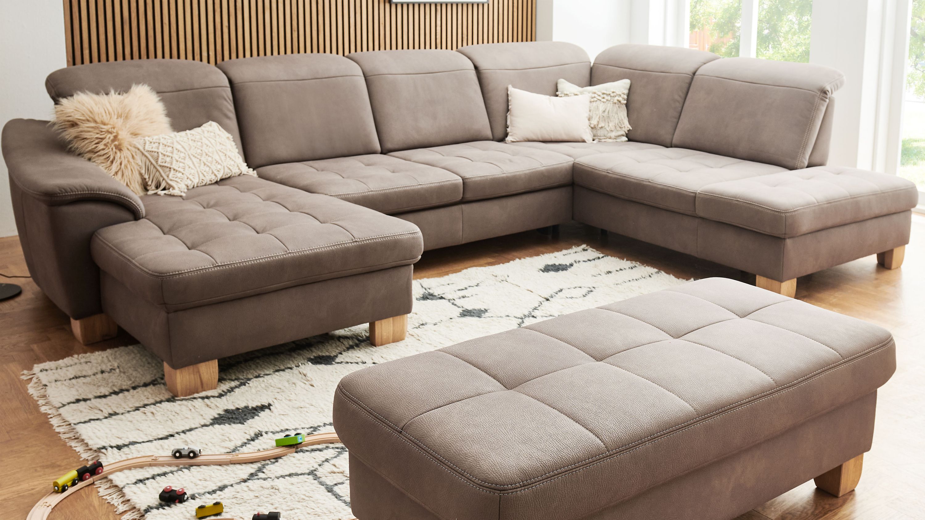 Wohnlandschaft mit eckigem Sofa und Hocker in braunem Stoff mit Holzfüßen in hellem Raum