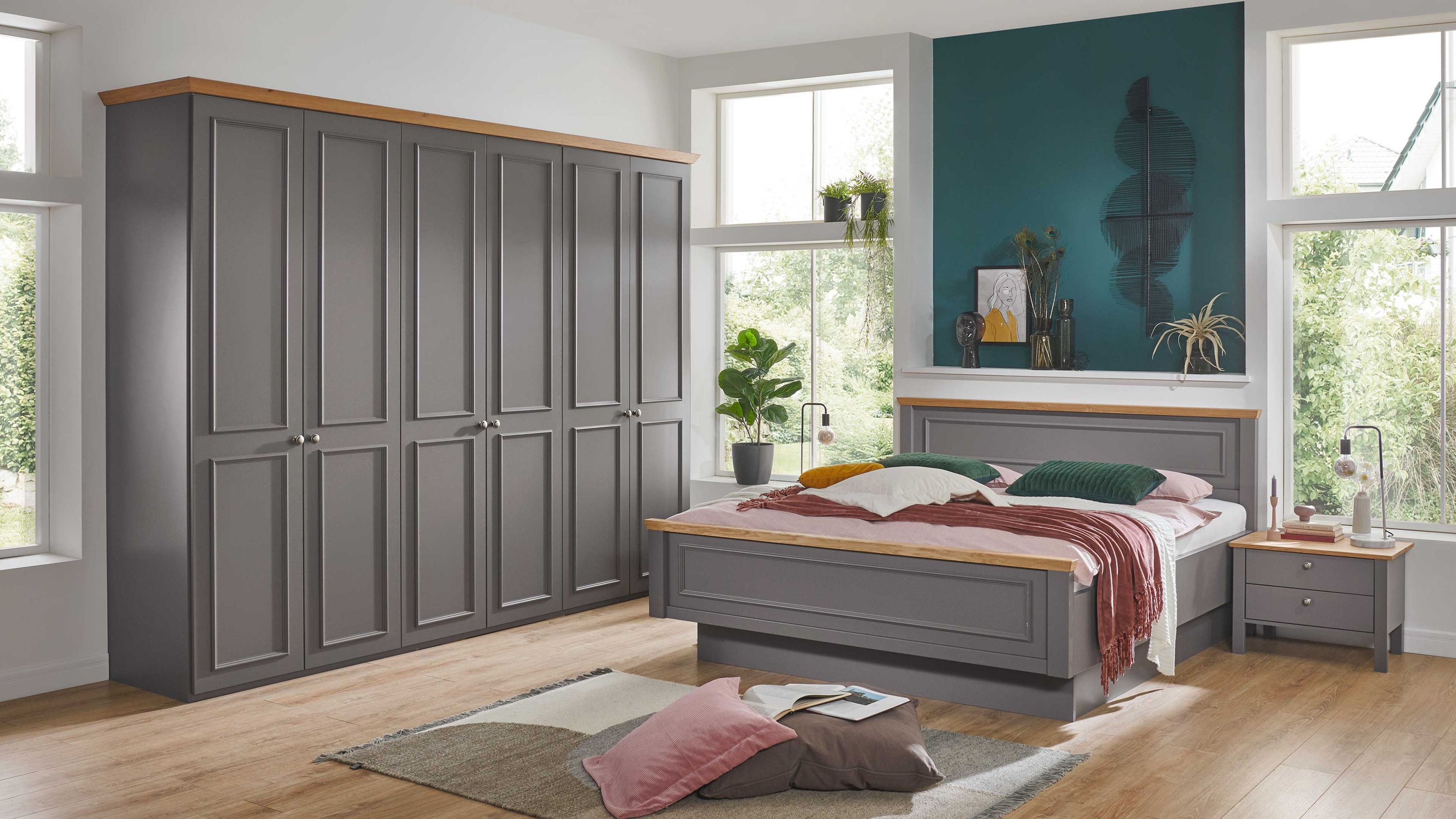 Schlafzimmer mit grauem Kleiderschrank grauem Bett mit Holzabsetzungen und grauem Nachttisch vor großen Fenstern