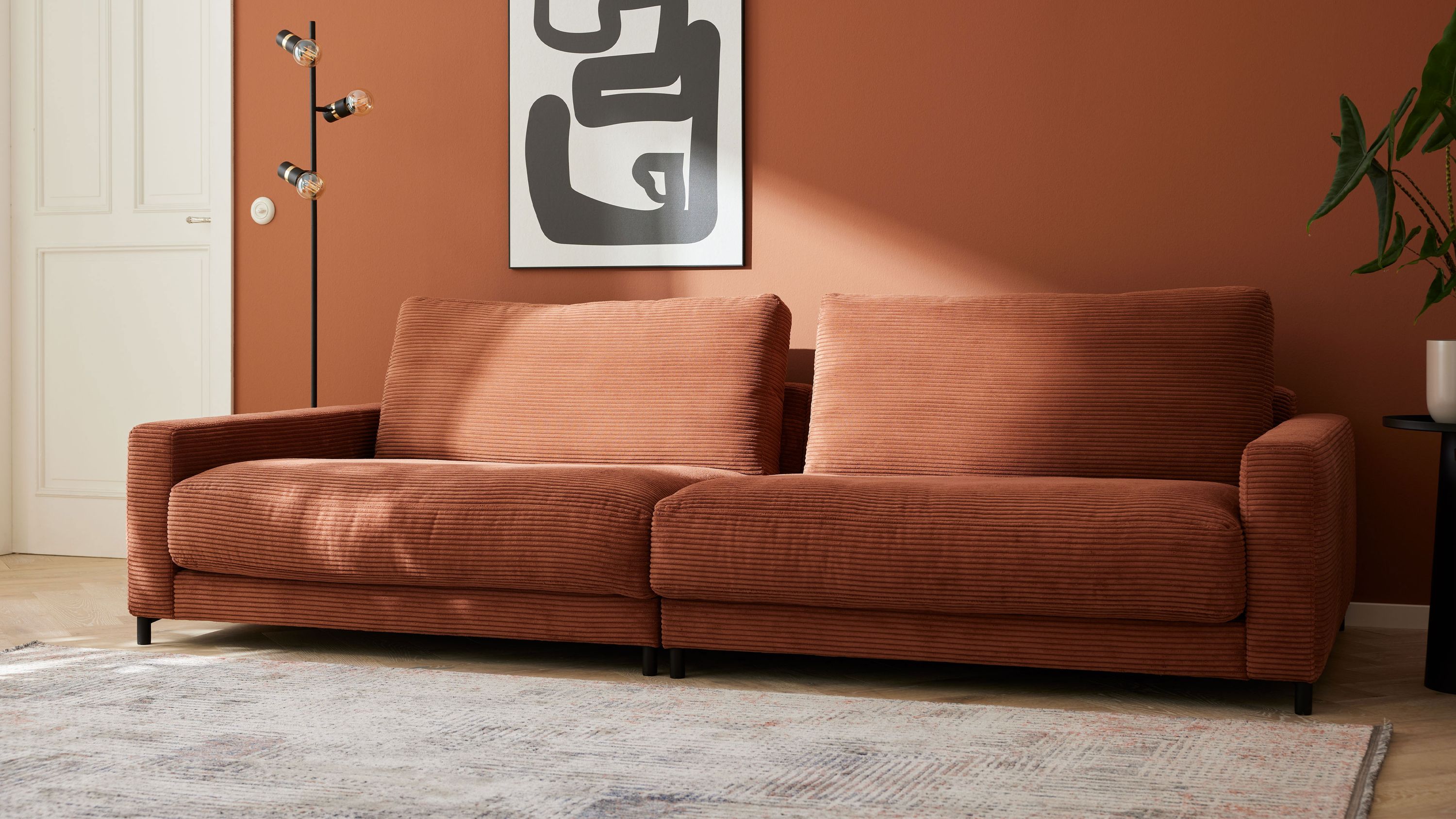 Zweisitziges Sofa mit rostfarbenem Cordbezug und schwarzen Metallfüßen vor rostfarbener Wand