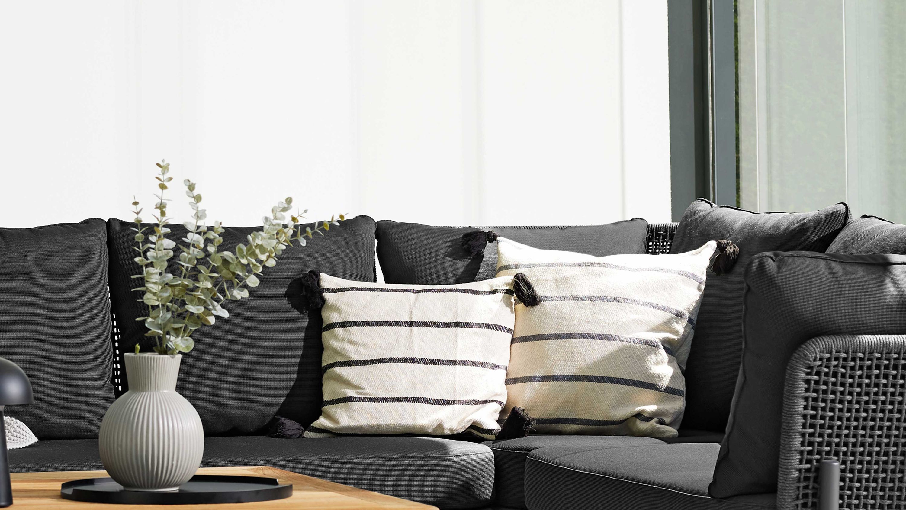 Eckmodul eines Gartensofas mit grauem Kordelgeflecht und dunkelgrauen Polstern mit zwei gestreiften Kissen