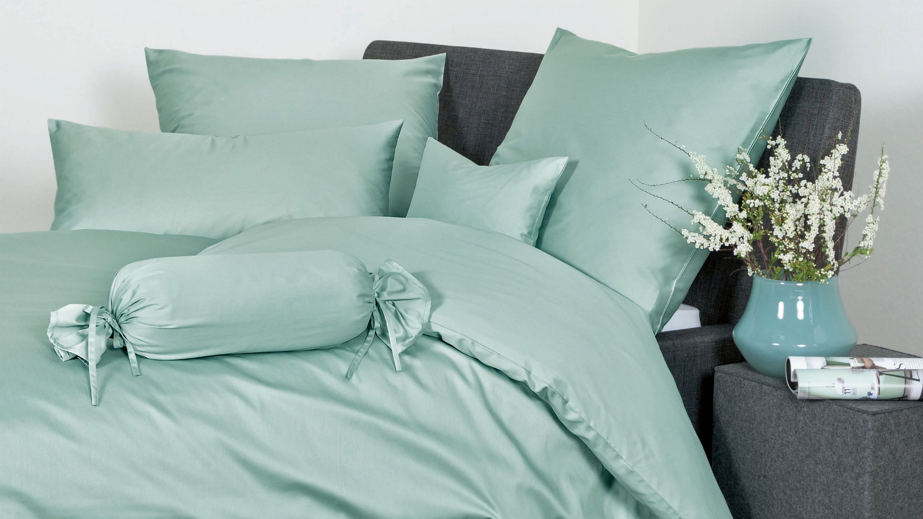 Hellgrüne Bettwäsche mit Kissen und Zierrolle auf dunklem Bett mit grauem Kopfteil und Nachttisch mit Vase und Blumen