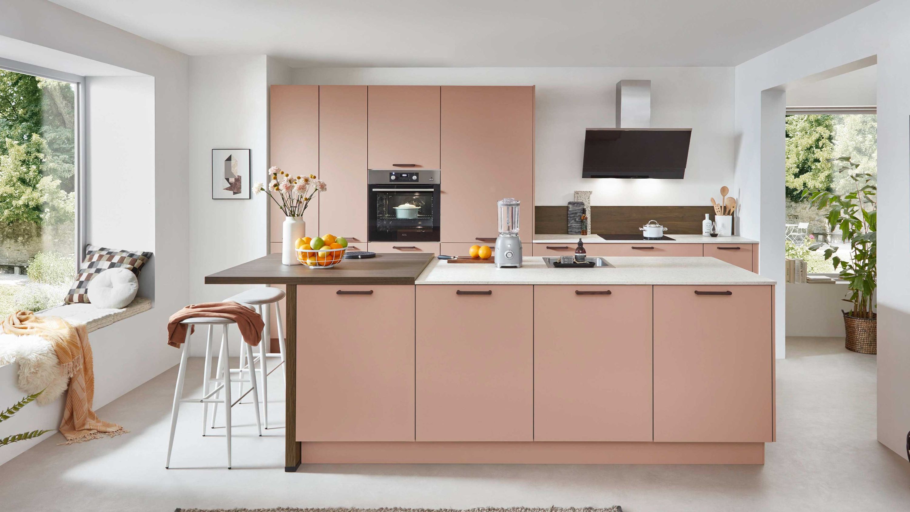 Inselküche in Rosé mit Holztheke und Einbauofen. Moderne Inselküche in Rosé mit glatten Fronten Holztheke Einbauofen und großer Fensterfront im Hintergrund.