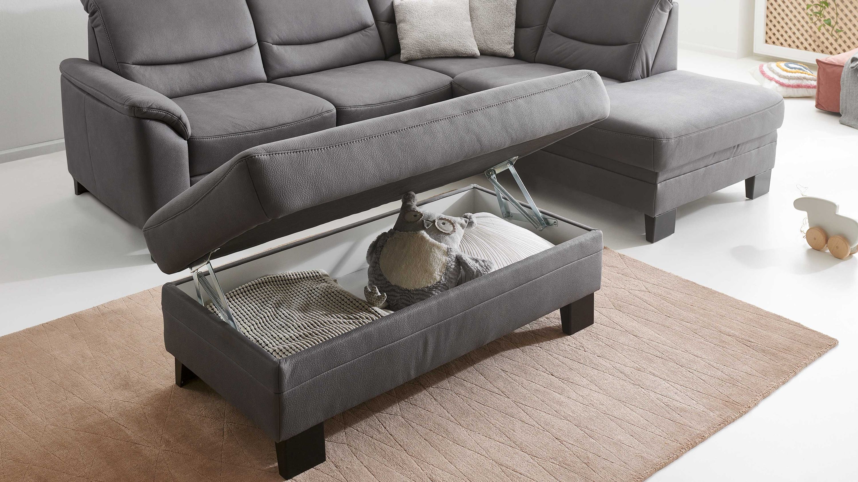 Grauer rechteckiger Hocker mit aufklappbarem Sitz und Stauraum schwarze Füße vor grauem Sofa