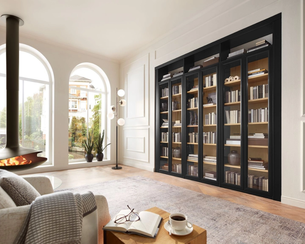 Stilvolles Wohnzimmer mit Kamin und moderner Bücherwand. Helles Wohnzimmer mit runder Fensterfront, Kamin, Bücherwand mit Glasfront und beiger Couchgarnitur auf Teppich.