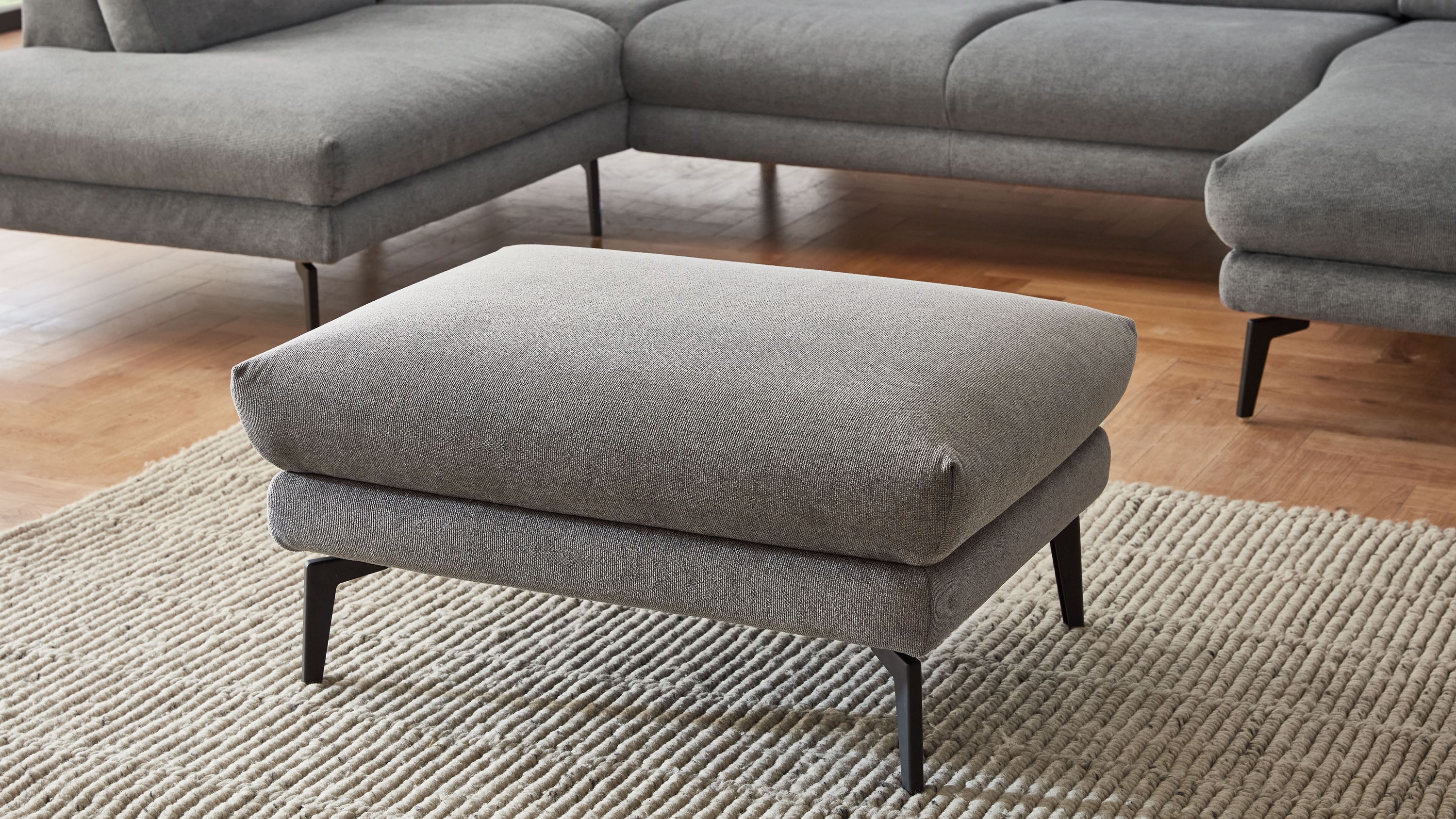 Quadratischer Hocker mit grauem Stoffbezug und schwarzen Metallfüßen auf hellem Teppich vor grauem Sofa
