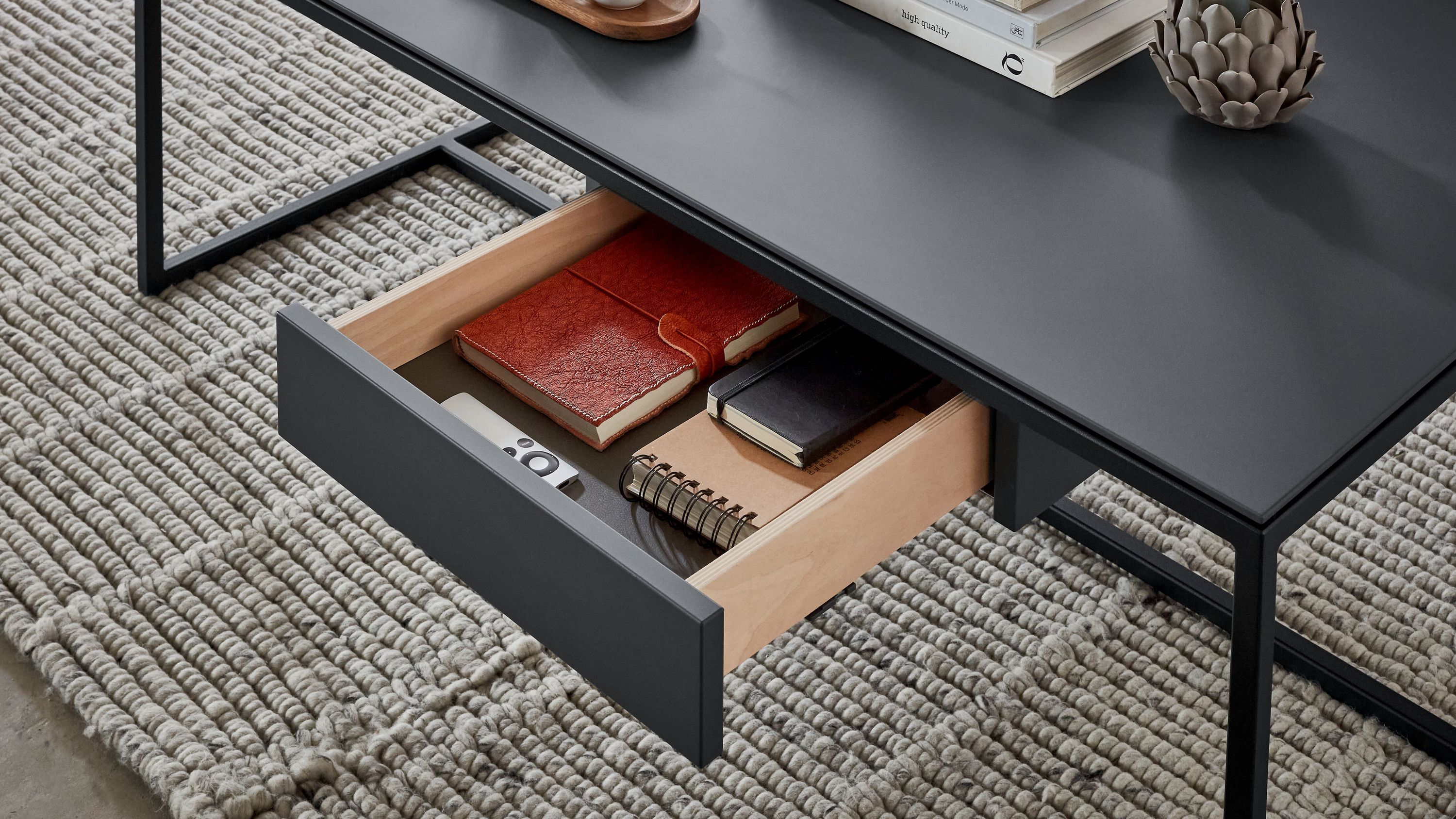 Offene Schublade eines graphitgrauen Tisches mit Büchern und Fernbedienung auf einem grauen Teppich