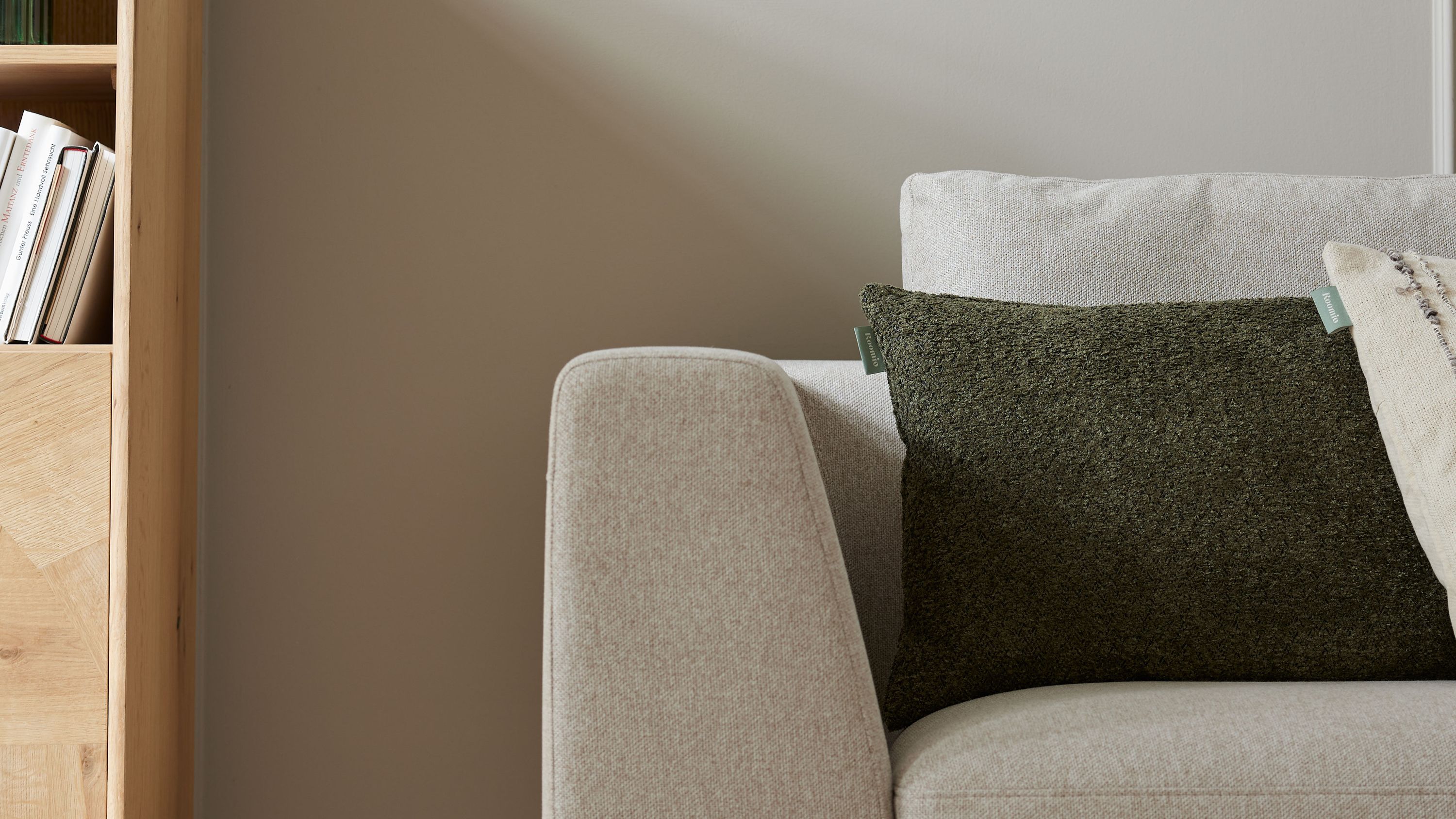 Rechteckiges Kissen in khakifarbenem Bouclé auf hellgrauem Sofa neben Holzregal mit Büchern