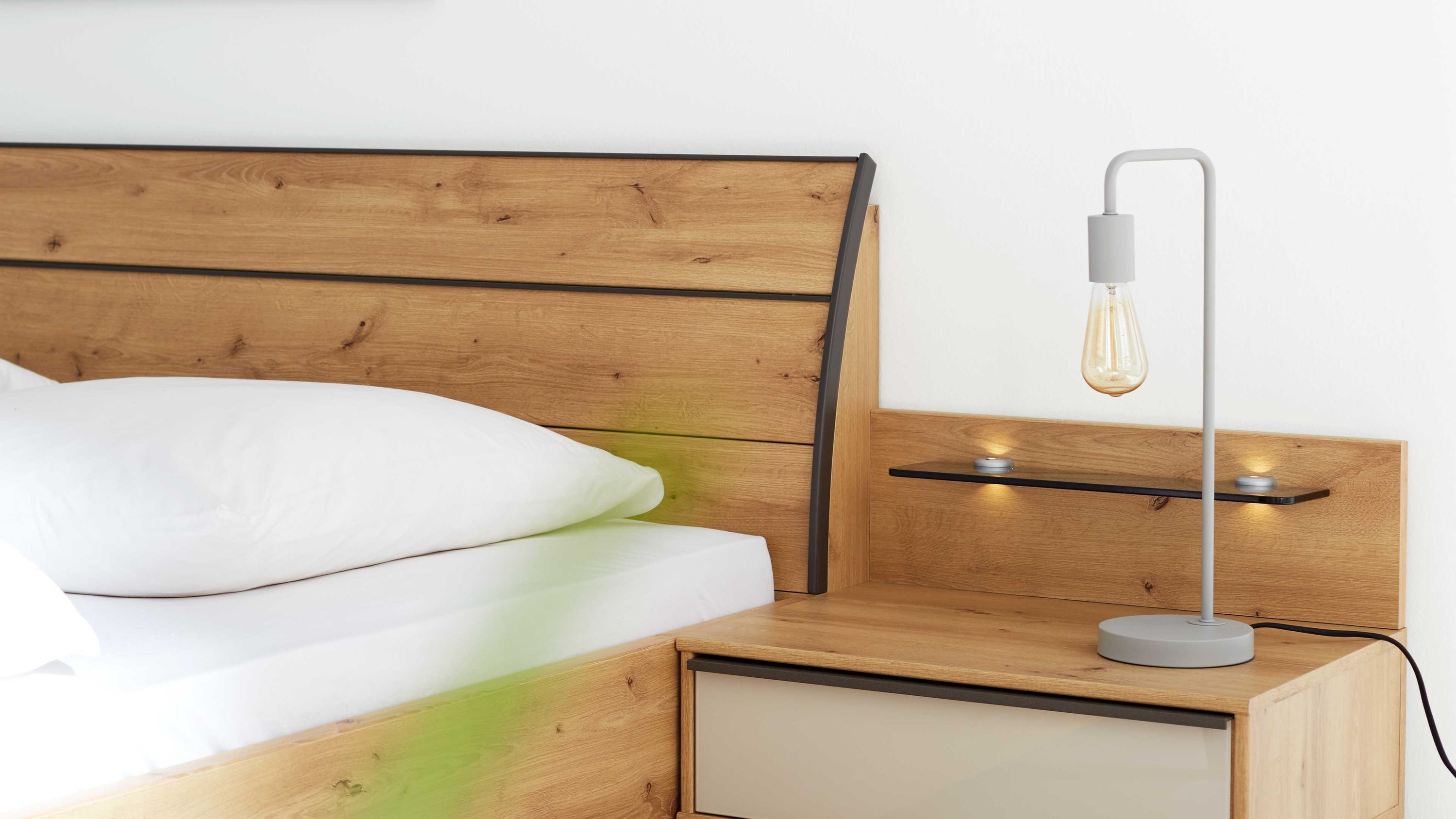 Holzbettrahmen mit Kopfteil aus hellbraunem Holz und schwarzem Streifen neben Nachttisch mit grauer Lampe