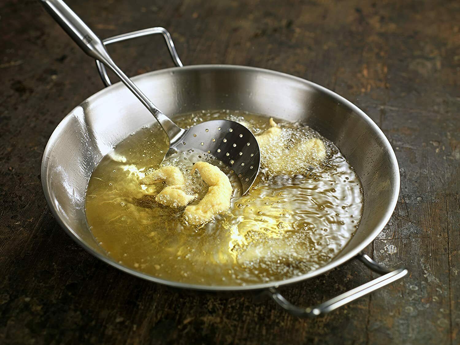 Metallpfanne mit Griffen, gefüllt mit heißem Öl, Schaumlöffel hält frittiertes Stück, frontal fotografiert.