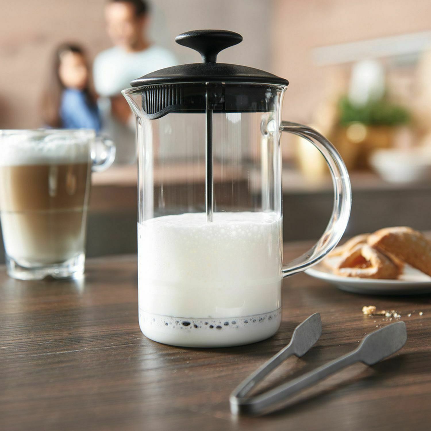 Milchaufschäumer mit Milchschaum auf Tisch, dahinter Kaffeetasse und Brot, aus leicht erhöhter Perspektive.