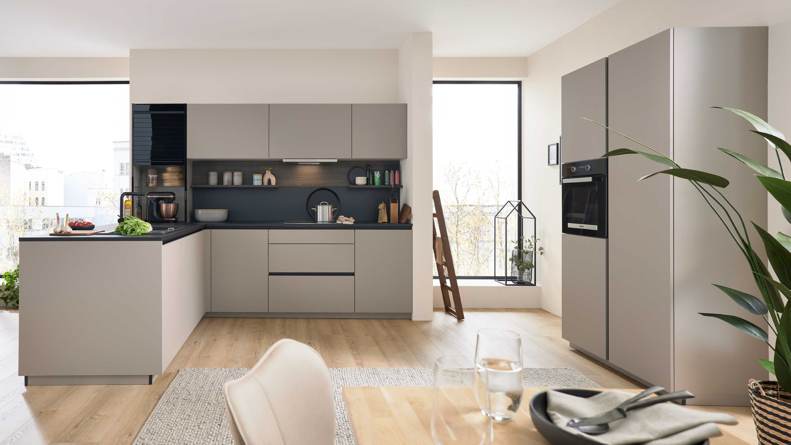 Küche mit L-Form und heller Einrichtung Helle Schränke L-Küche mit Einbaugeräten Fensterlicht gedeckter Esstisch Pflanzen und Regaldeko