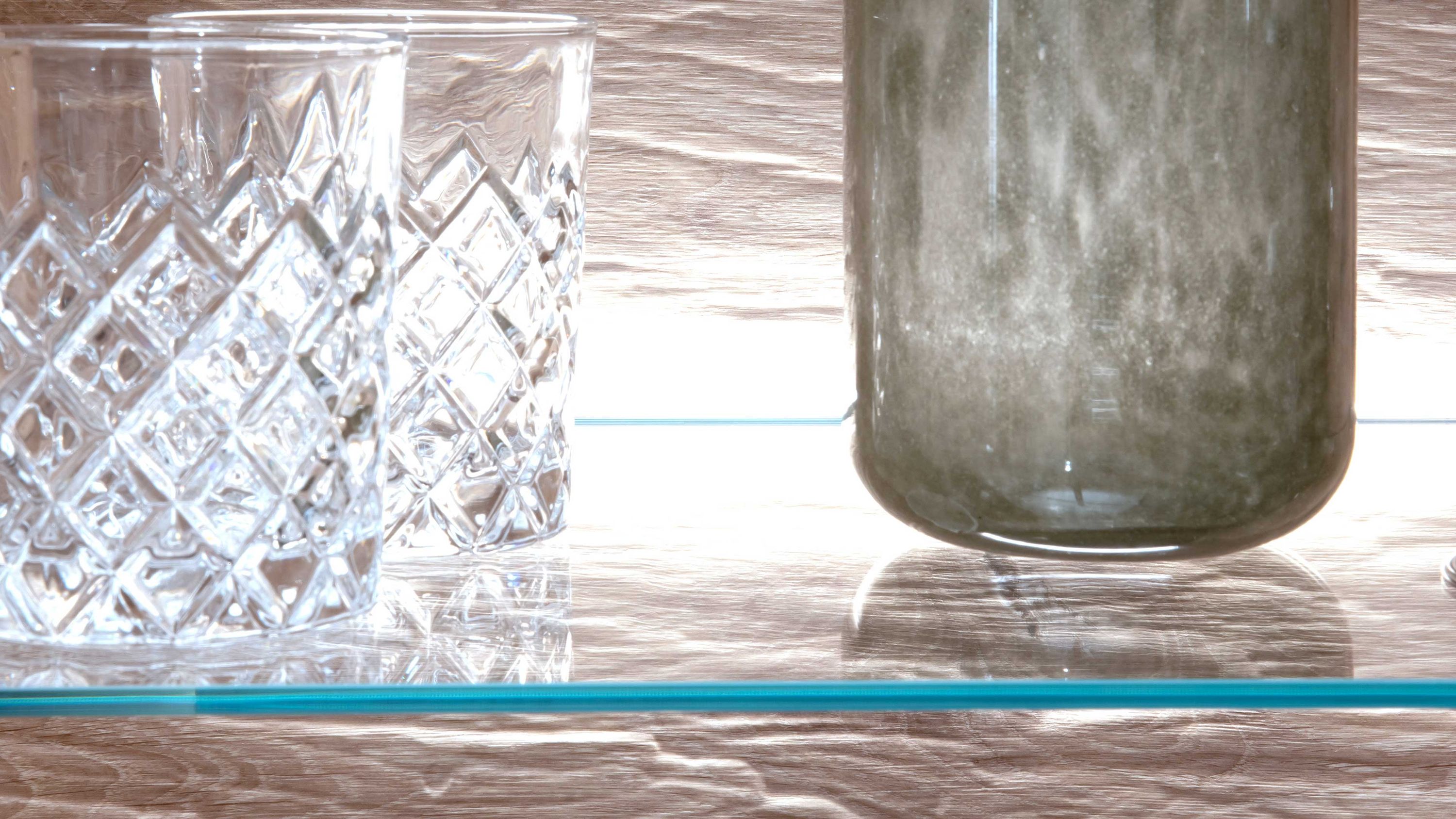 Glasregal mit zwei transparenten Gläsern mit Muster und einer grauen Glasvase auf Holzrückwand mit Beleuchtung
