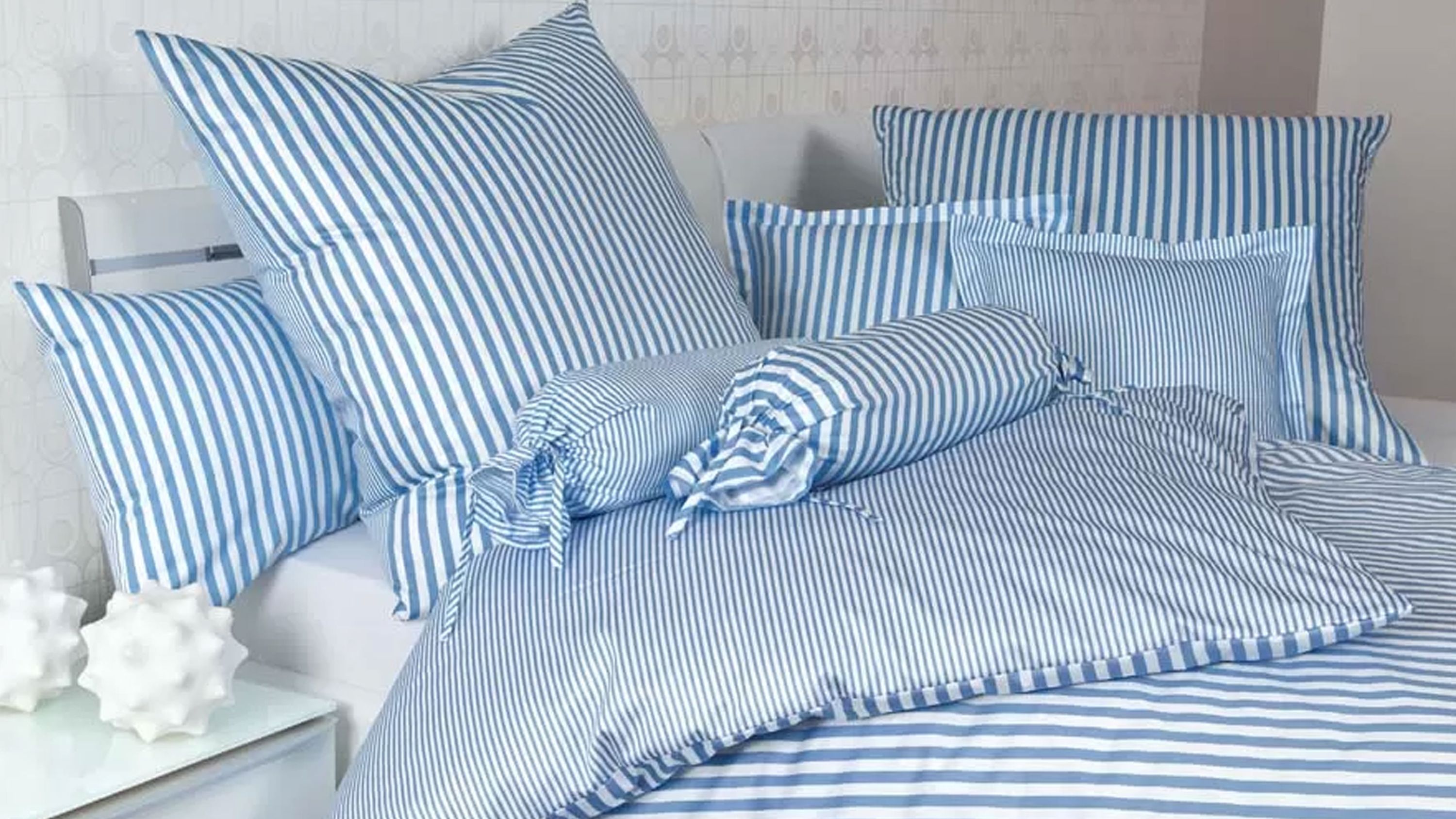 Hellblaue Bettwäsche mit weißen Streifen auf Bett mit mehreren Kissen und Rollenpolstern