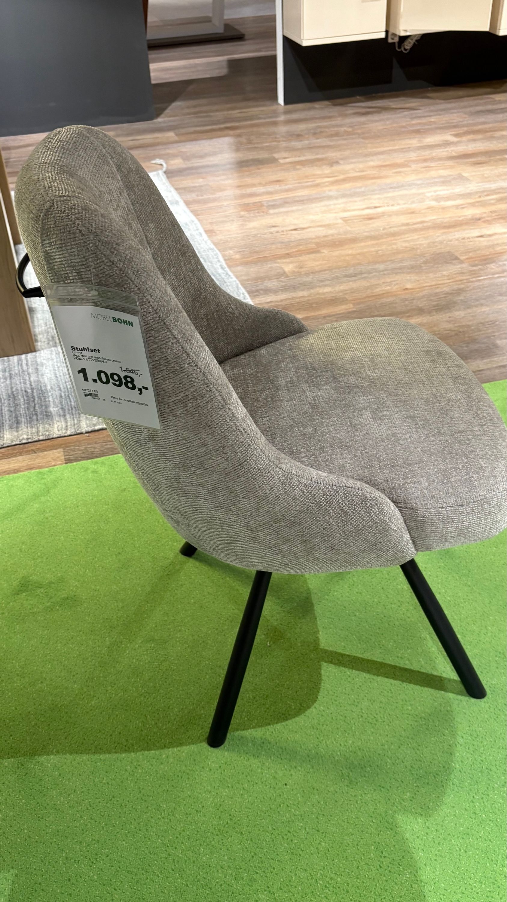 Grauer Stuhl mit abgerundeter Rückenlehne, schwarzem Gestell und Preisschild auf grünem Teppichboden in Ausstellung