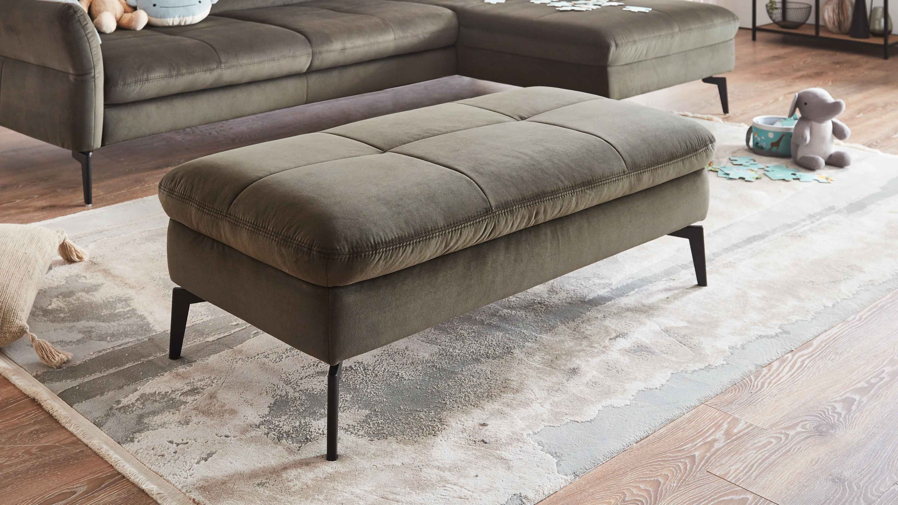 Großer rechteckiger Hocker mit olivgrünem Bezug und schwarzen Metallfüßen auf hellem Teppich vor Sofa