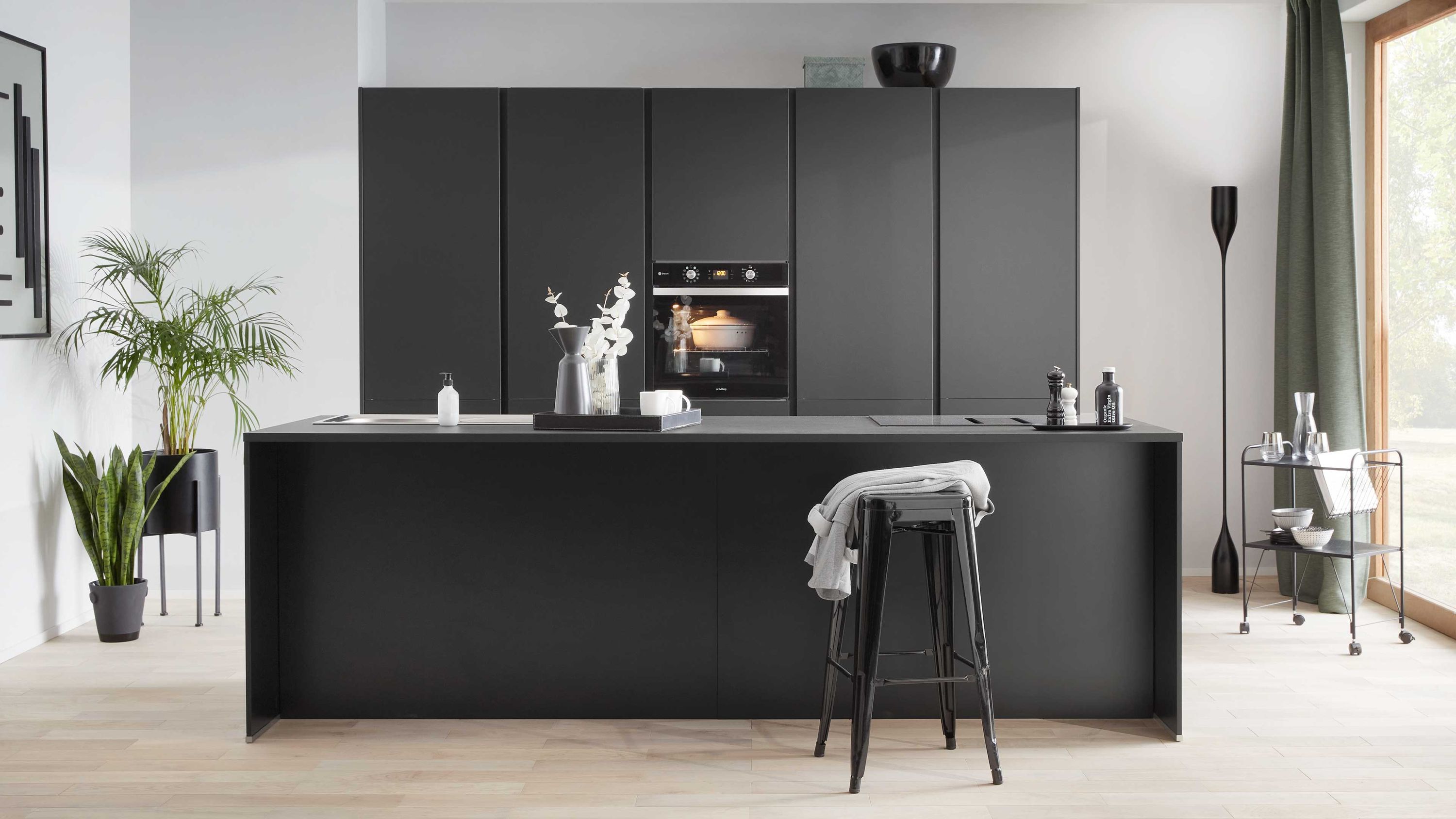 Schwarze Küche mit Insel Holzfußboden Einbauofen Barhocker mit Tuch Pflanze Fenster mit grünen Vorhängen.