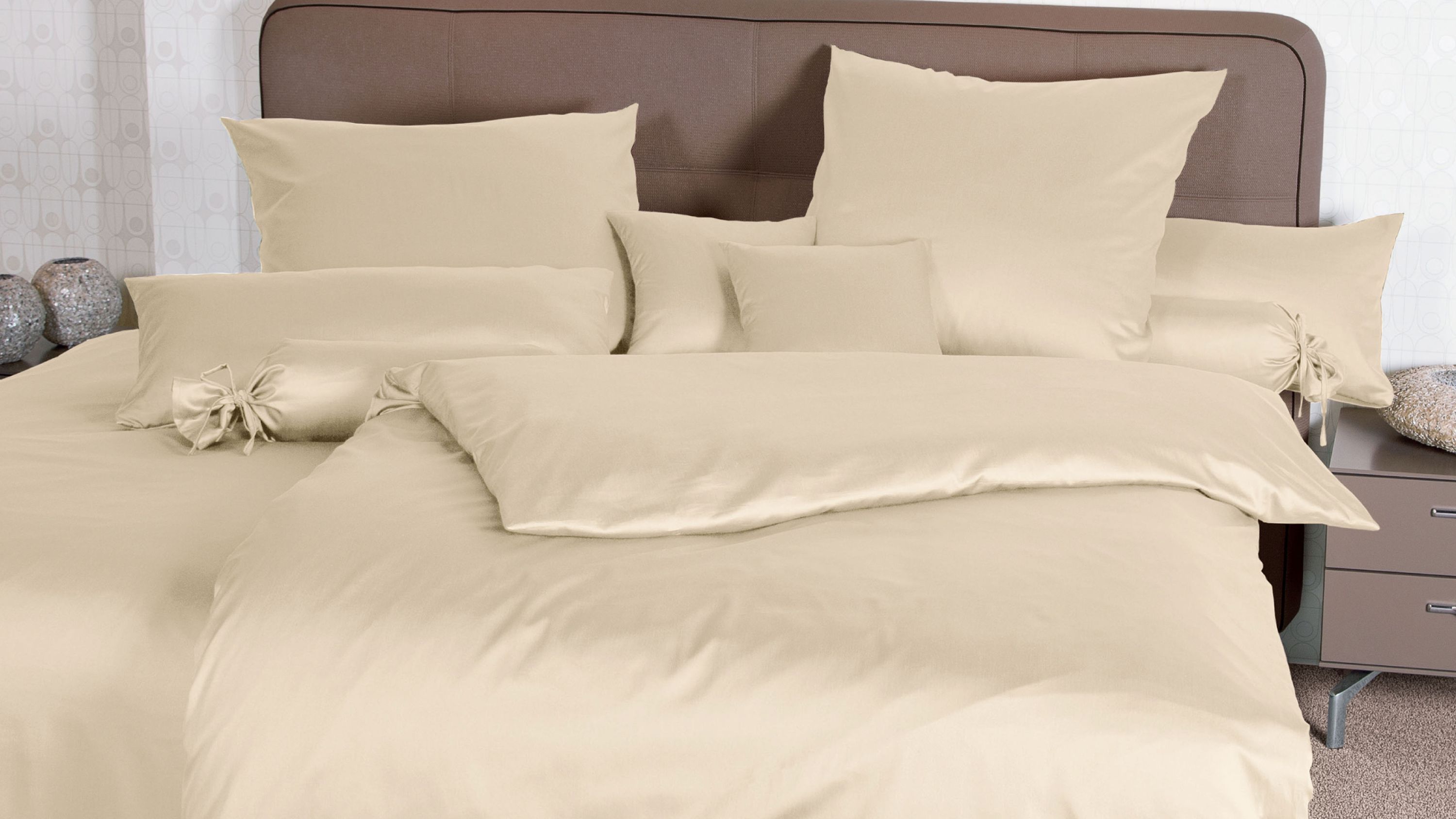 Bett mit cremefarbener Bettwäsche bestehend aus Kissen Zierkissen und Decke vor braunem Kopfteil