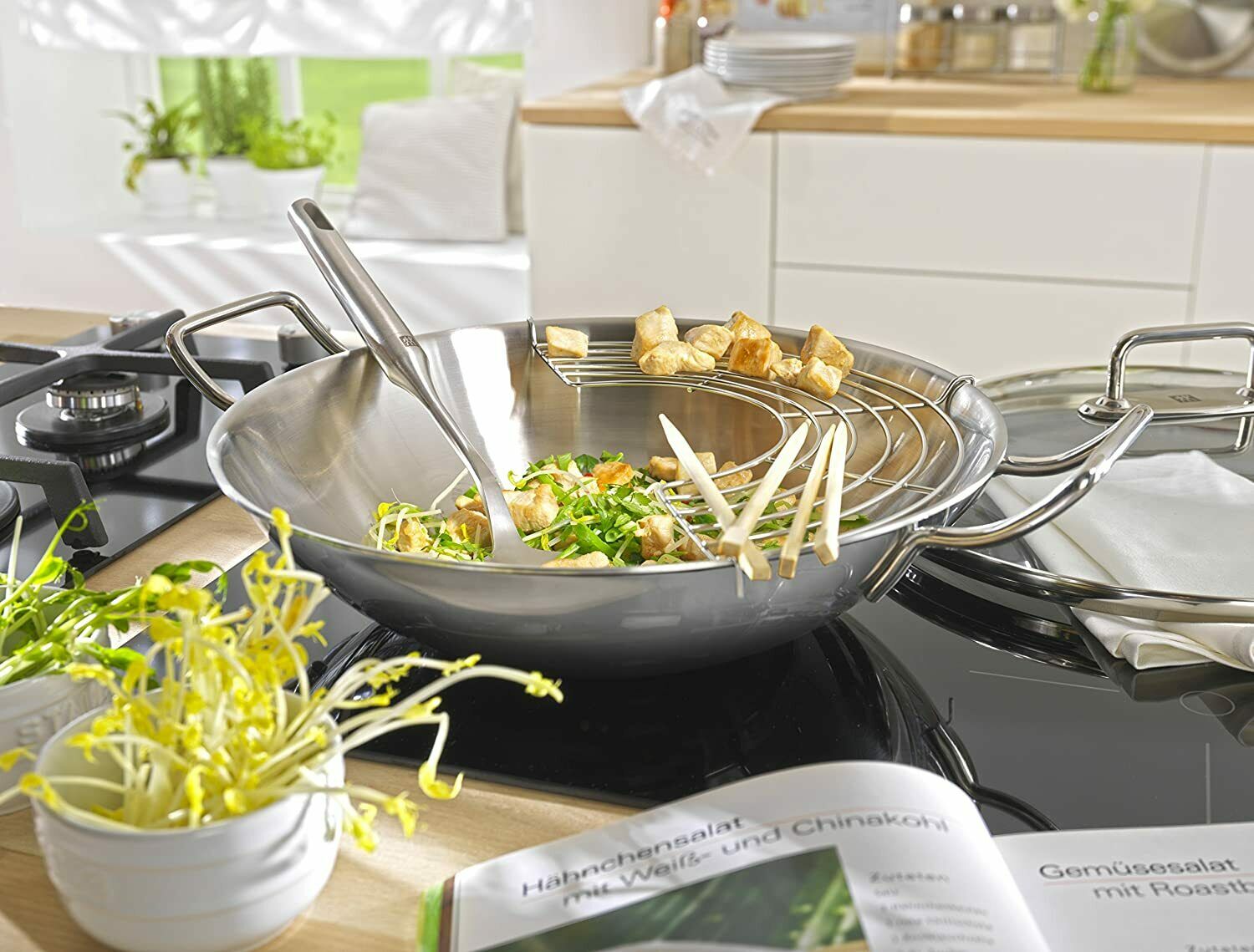 Wok mit Salat auf Küchentheke, umgeben von Küchenutensilien, Zutaten und Deko, leicht schräg fotografiert.