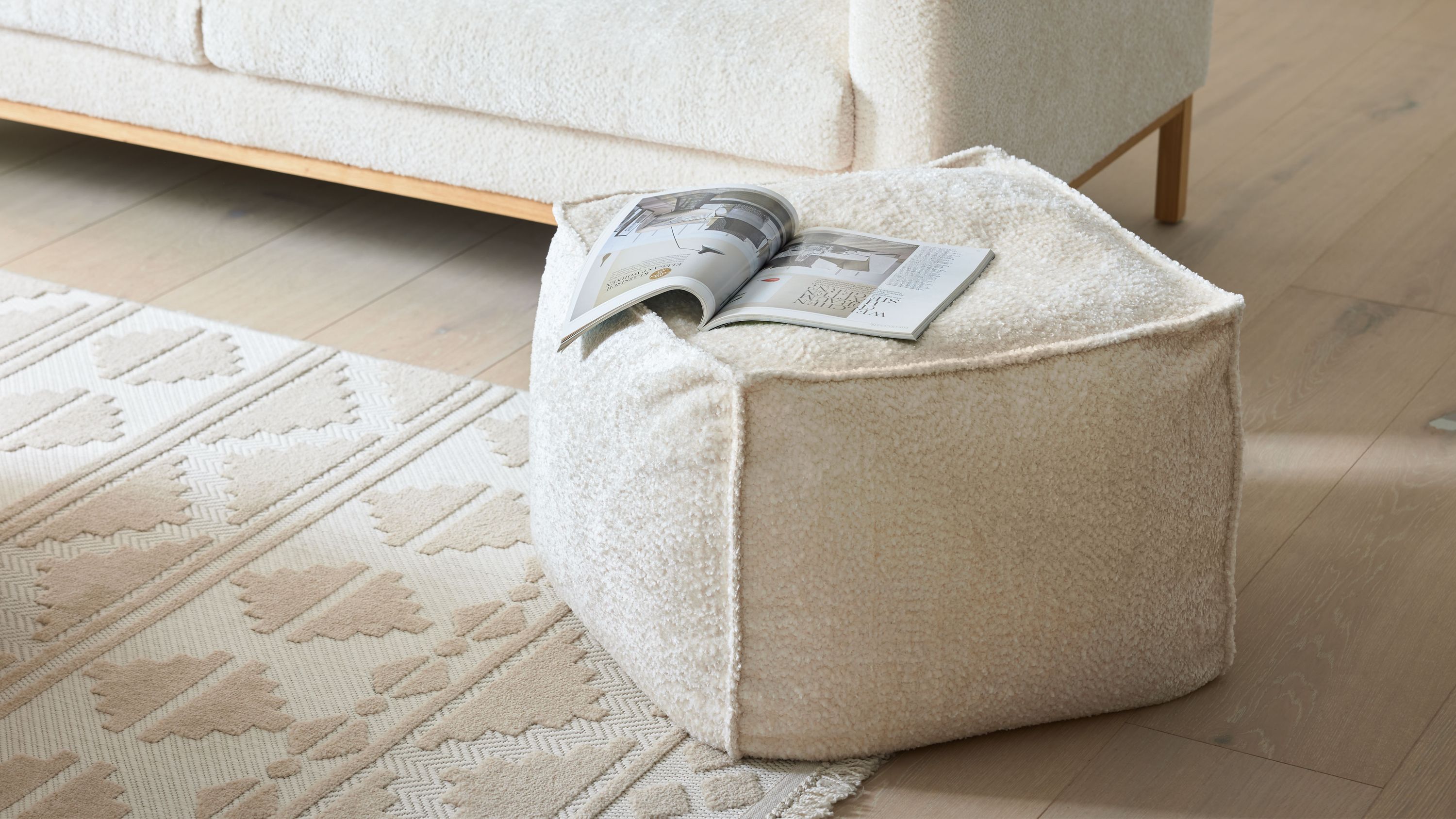 Quadratischer Hocker mit weißem Stoffbezug auf hellem Holzboden neben Sofa und Teppich