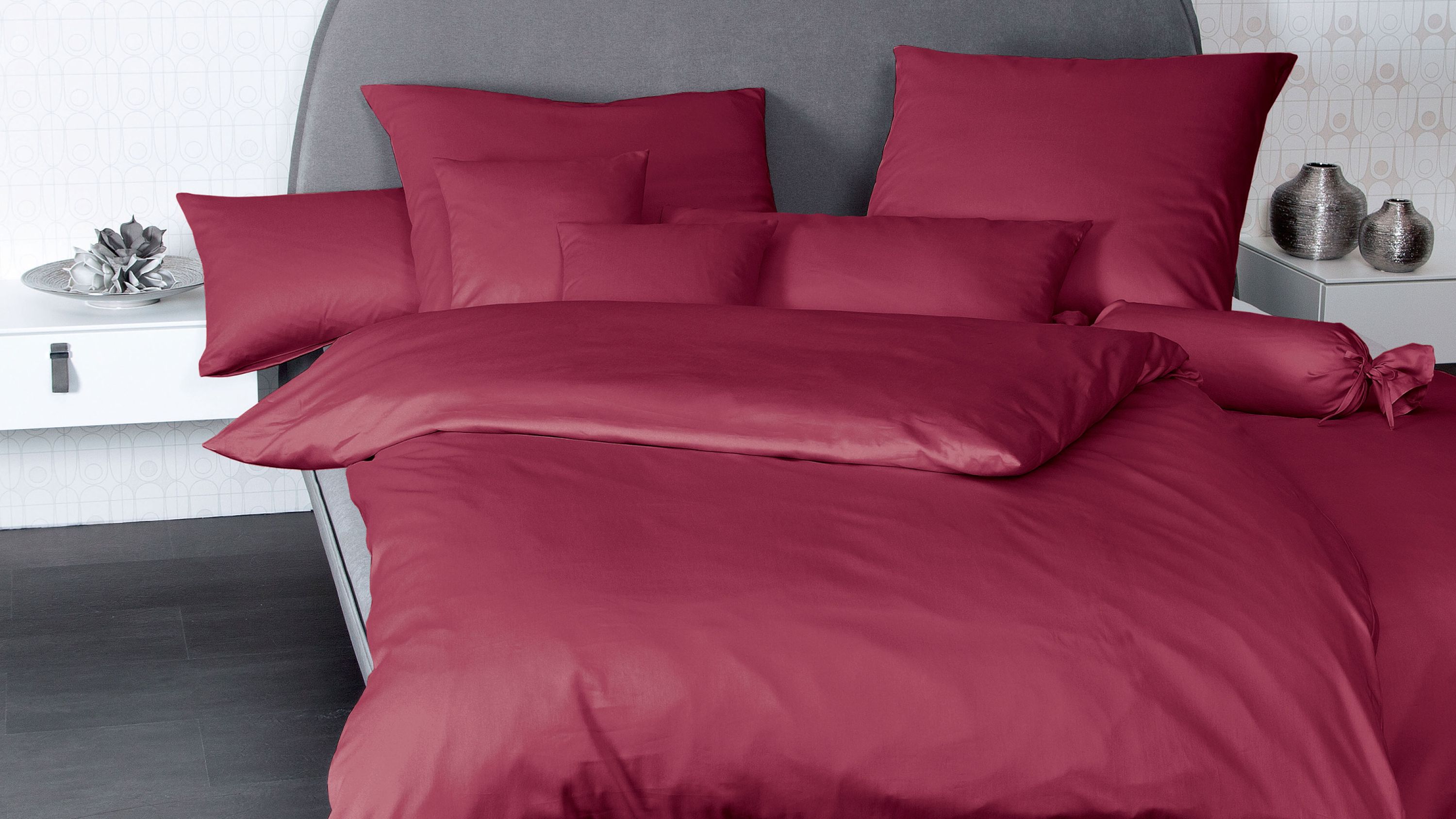 Bett mit bordeauxroter Bettwäsche bestehend aus Kissen und Decke vor grauem Kopfteil
