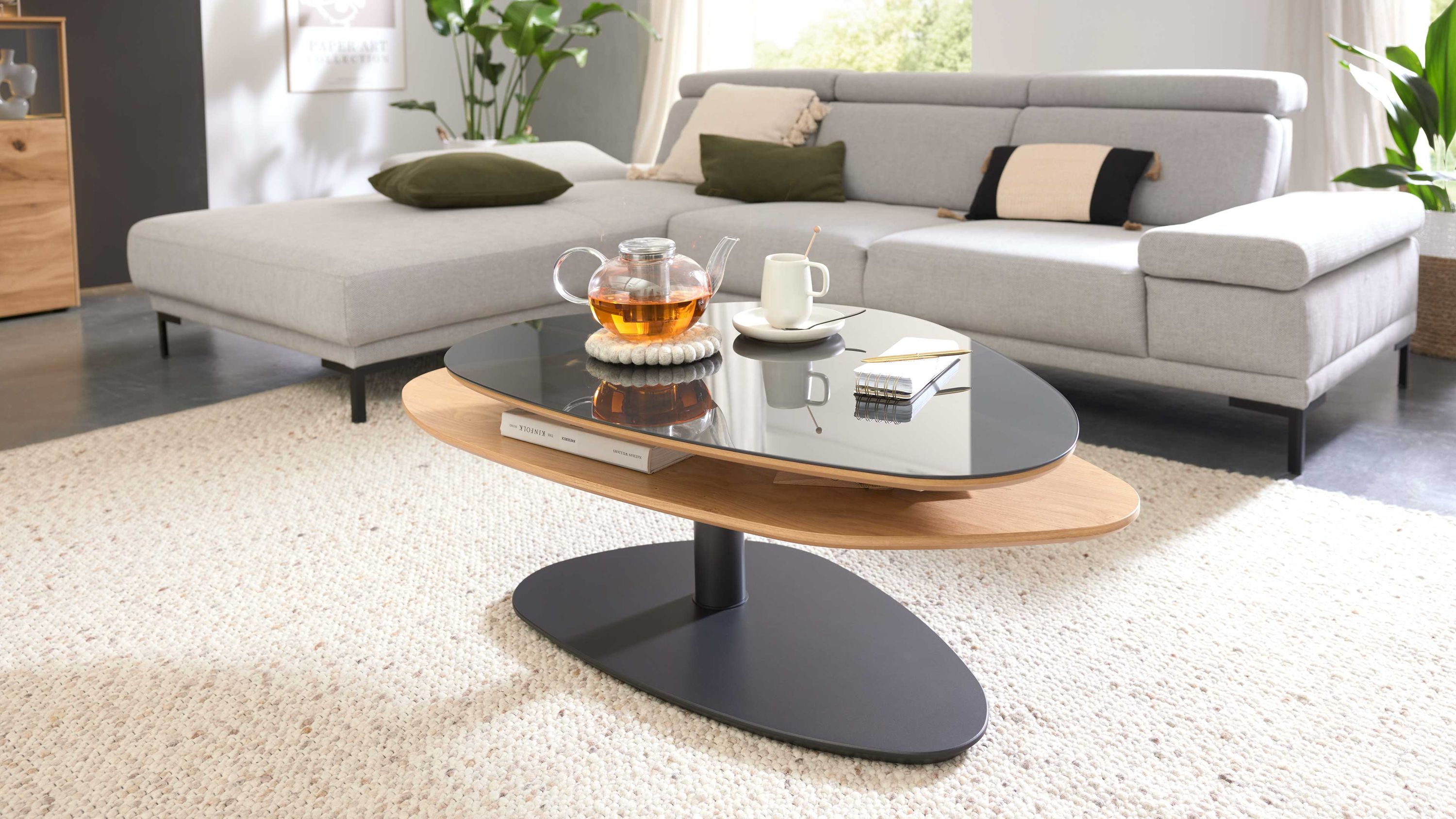 Ovaler Couchtisch mit schwarzer Glasplatte und Holzablage auf schwarzem Sockel vor grauem Sofa