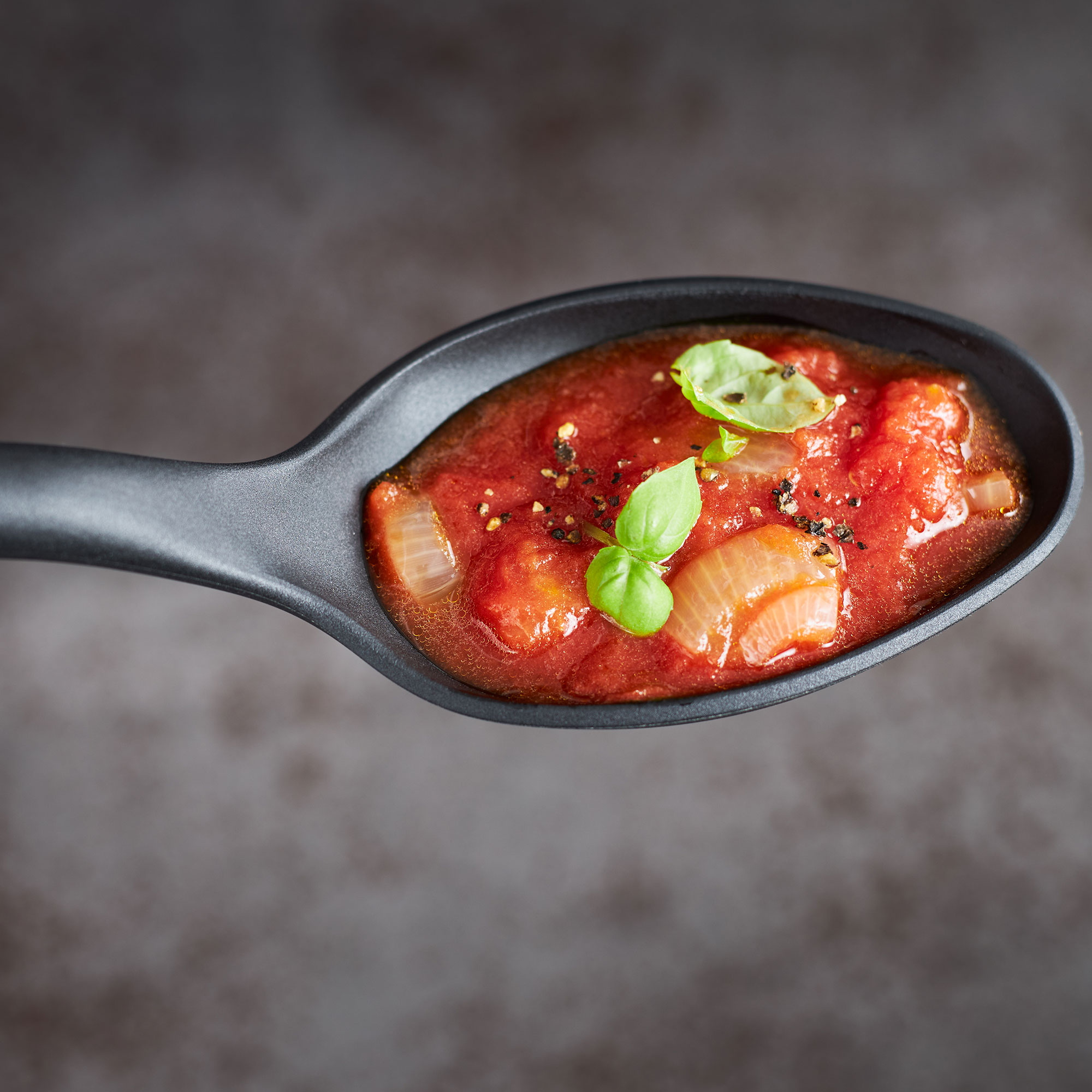 Nahaufnahme eines Schöpflöffels aus Edelstahl mit roter Sauce Gemüse & Kräutern schräg von oben unscharfer Hintergrund