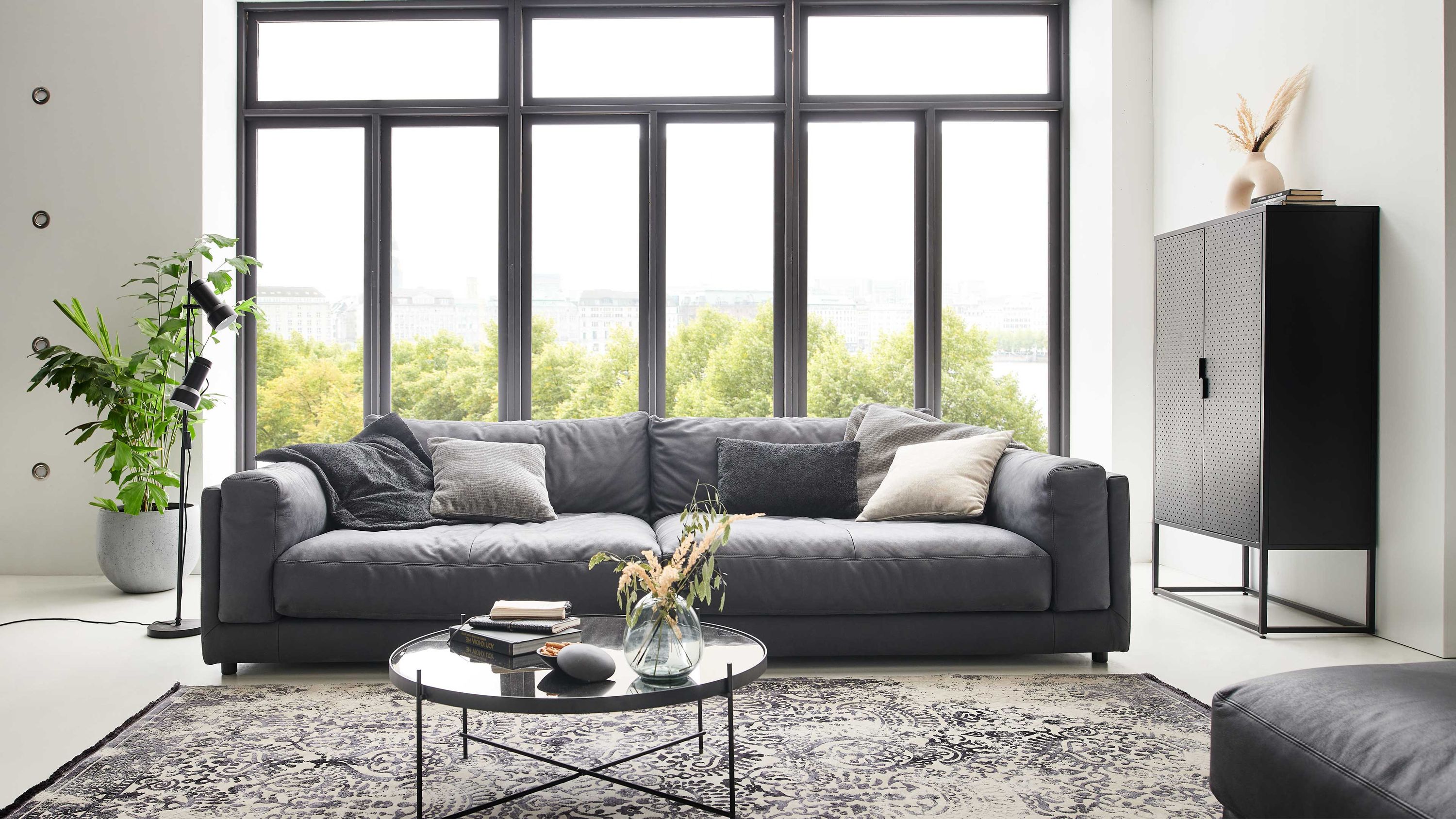 Großes anthrazitfarbenes Sofa mit Kissen vor großen Fenstern in modernem Wohnzimmer