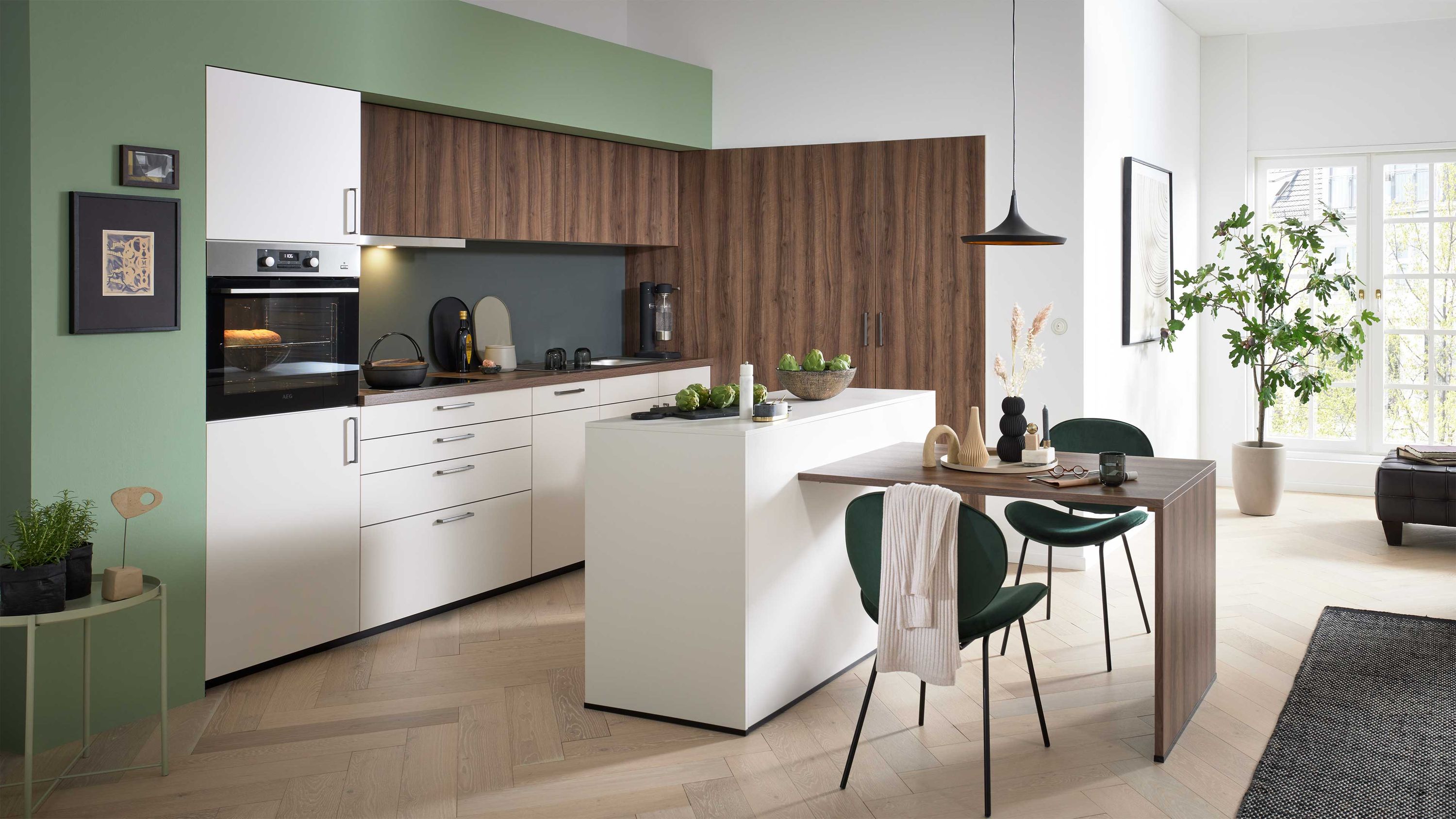 Helle Küche mit Essbereich und grünen Akzenten Weiße Küche mit Holzdetails schwarzer Rückwand Insel mit Tisch grünen Stühlen Hängelampe Pflanzen Tageslicht.