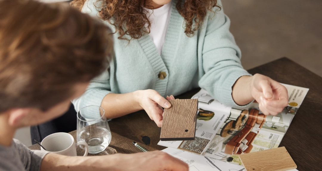 Zwei Personen vergleichen Holzproben über Grundrissen und Katalogen, Innenraumplanung und Materialauswahl für modernes Raumdesign