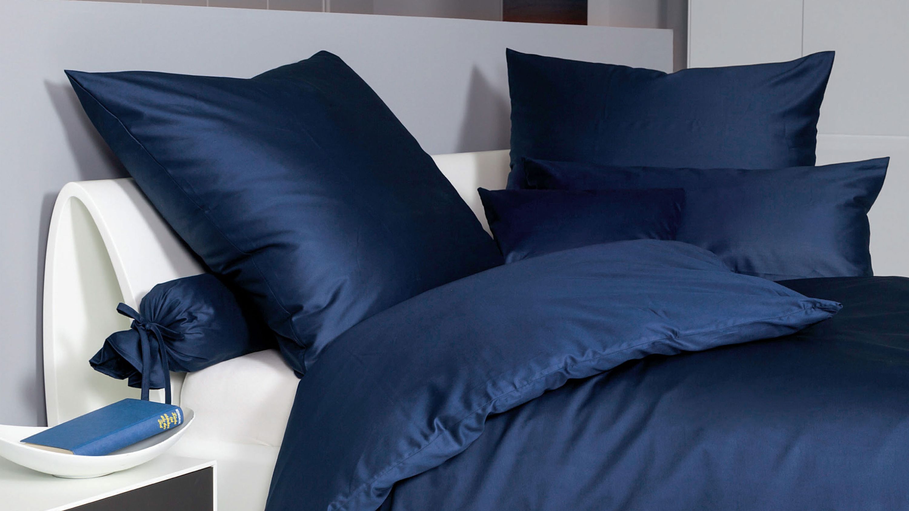 Dunkelblaue Bettwäsche mit mehreren Kissen auf weißem Bett mit geschwungenem Kopfteil