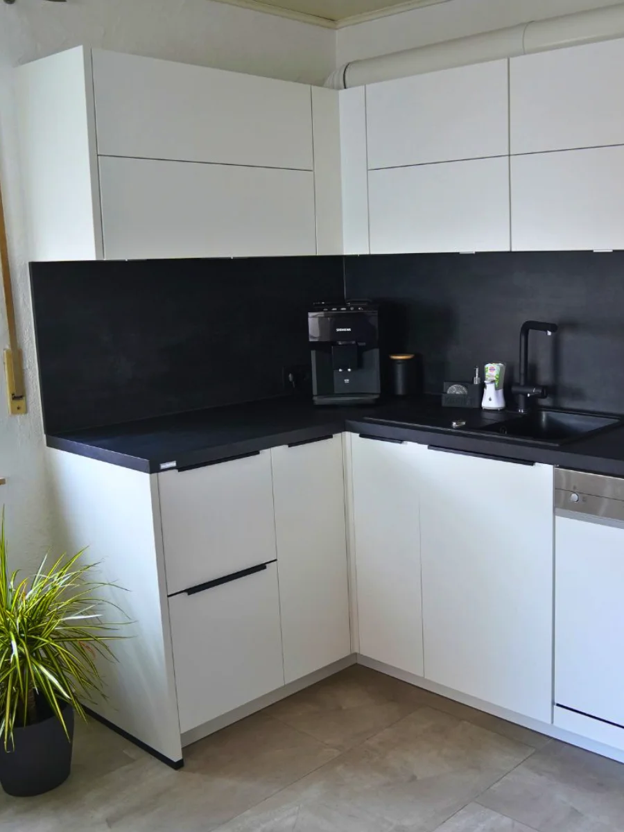 Moderne weiße Eckküche mit schwarzen Arbeitsflächen, Kaffeemaschine, Spüle, Einbaugeräten und Pflanze auf Fliesenboden.