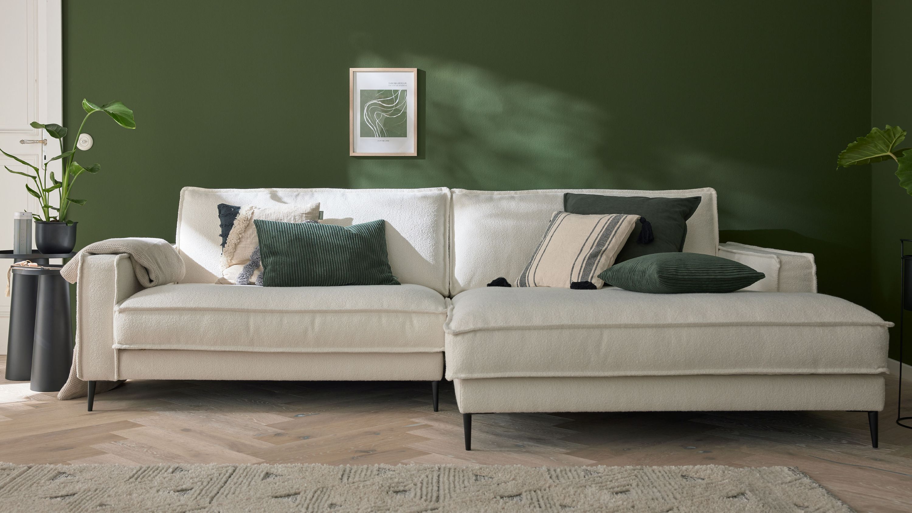 Ecksofa mit weißem Stoffbezug Metallfüßen und mehreren Kissen vor grüner Wand