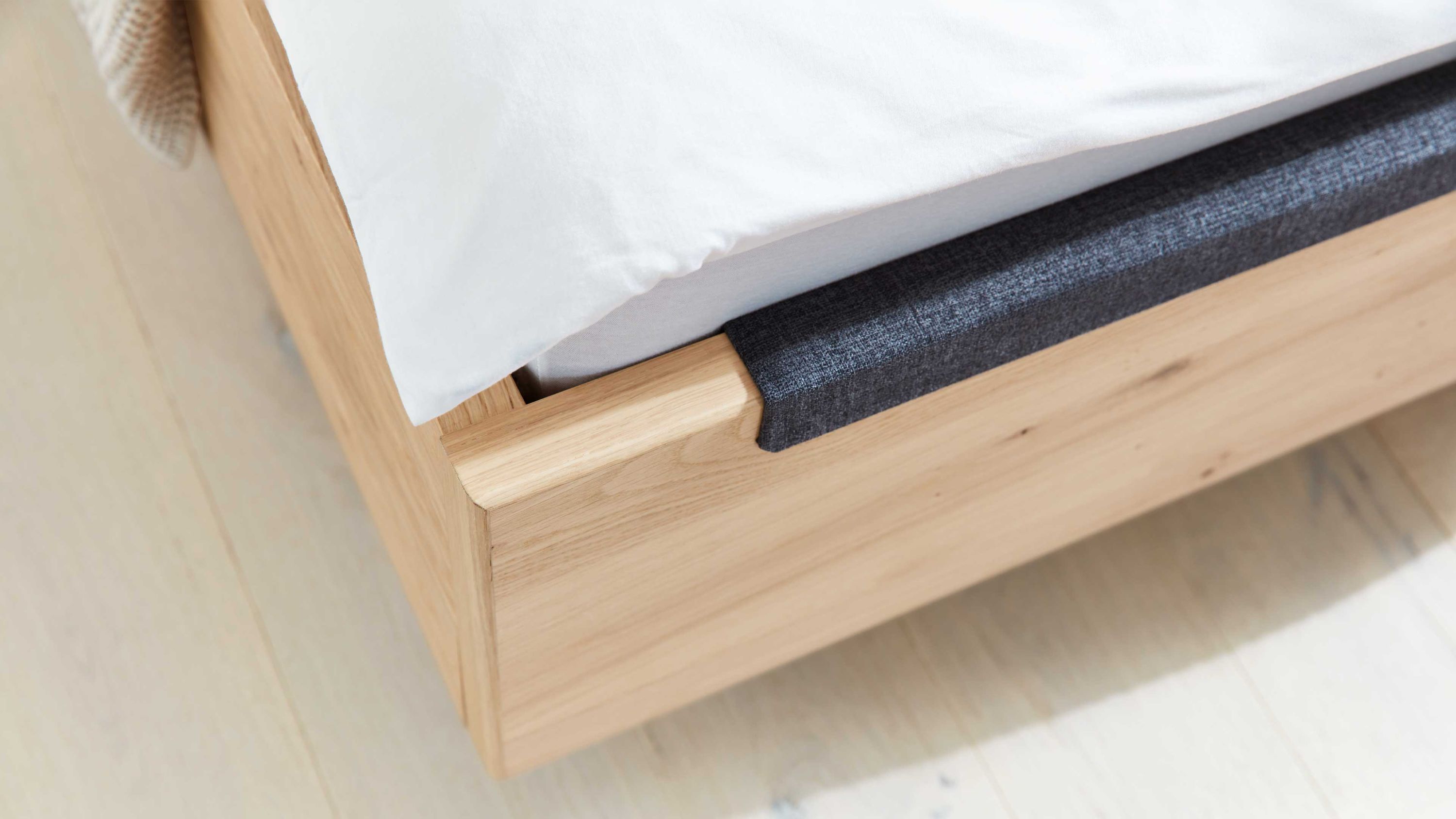 Bettrahmen mit anthrazitfarbener Polsterkante und hellem Holz Eckansicht eines Bettrahmens aus hellem Holz mit anthrazitfarbenem Polsterbezug an der Kante und weißer Bettwäsche