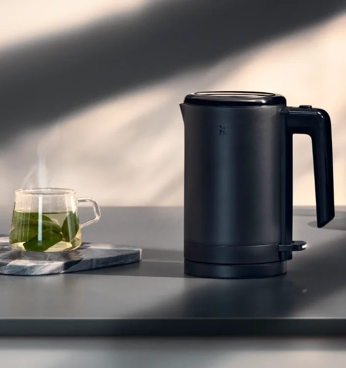 Wasserkocher WMF Schwarzer Wasserkocher von WMF neben einer dampfenden Tasse grünem Tee auf grauer Arbeitsplatte.