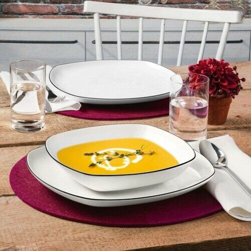 Teller mit Kürbissuppe auf weißem Porzellanservice  gedeckter Holztisch mit Deko, Besteck und weinroter Platzmatte