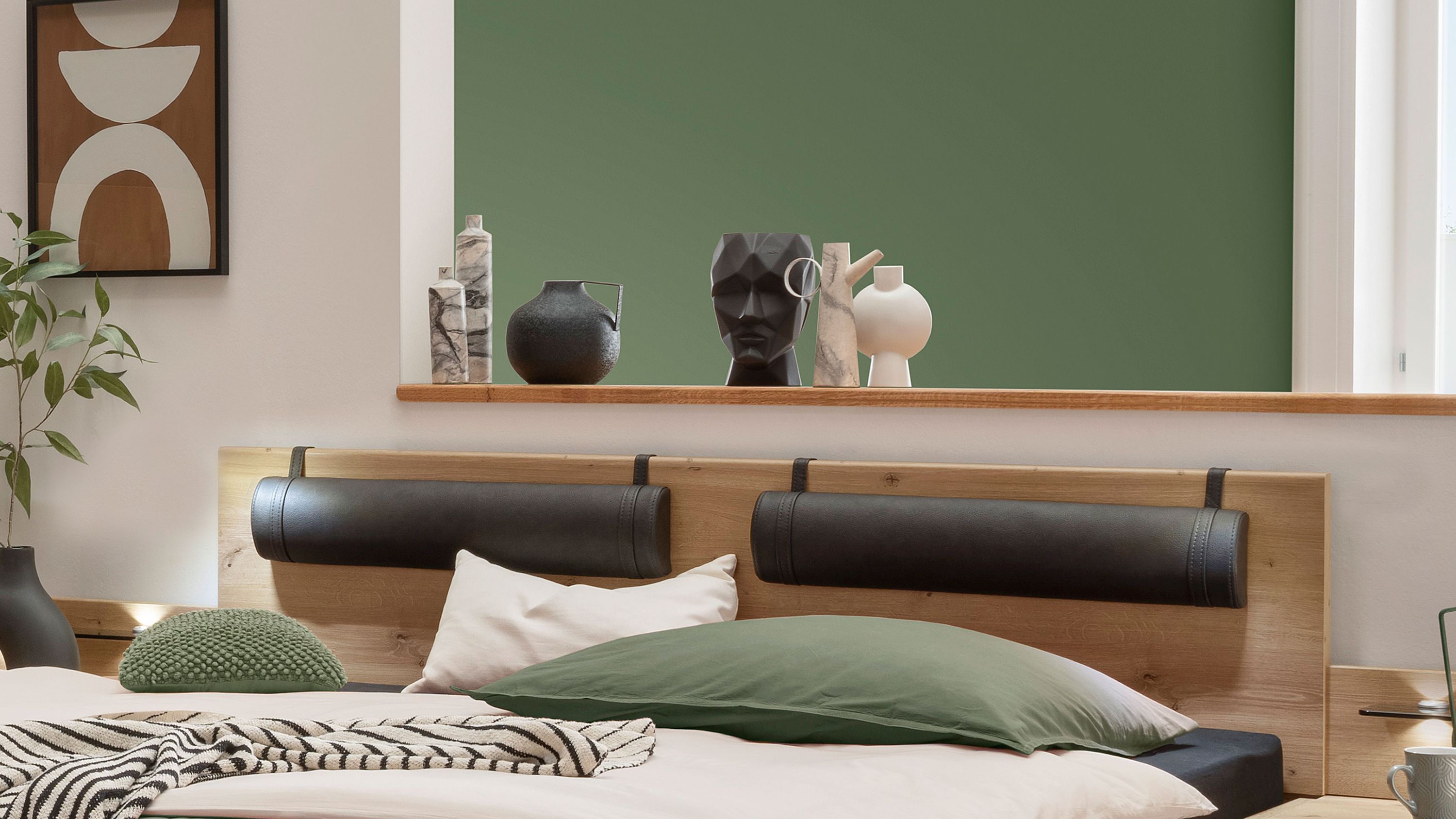 Holzbetthaupt mit zwei dunkelgrauen Polsterrollen aus Kunstleder davor weiße und grüne Kissen auf Bett