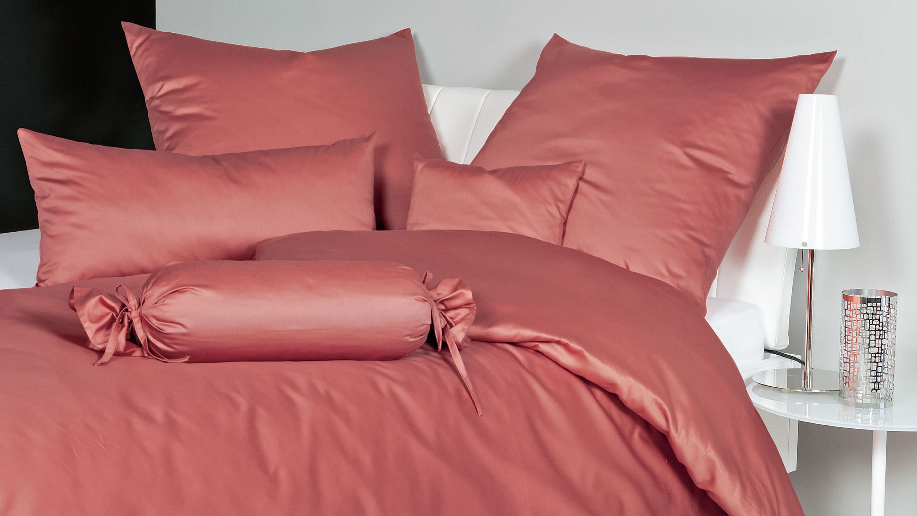 Rosa Bettwäsche-Set mit Kissen und Zierrolle auf weißem Bett mit Nachttisch und Lampe