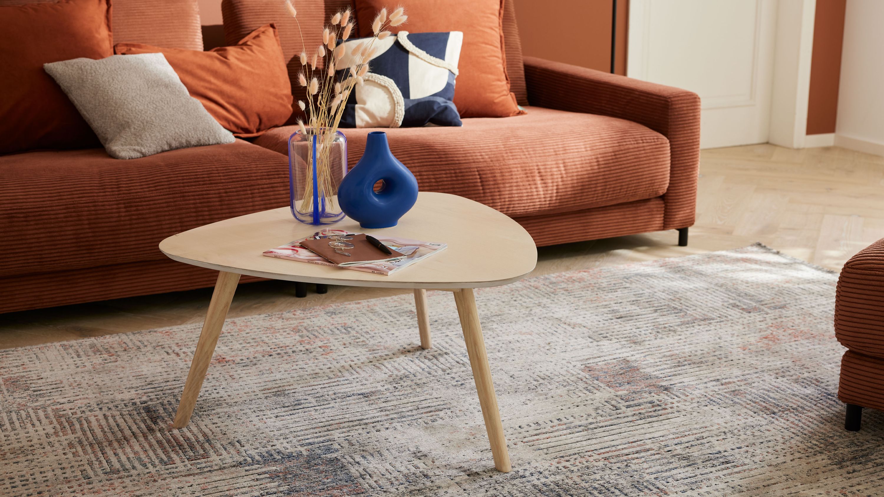Dreieckiger Couchtisch mit hellem Holz und drei Beinen darauf blaue Vasen und Zeitschriften vor braunem Sofa