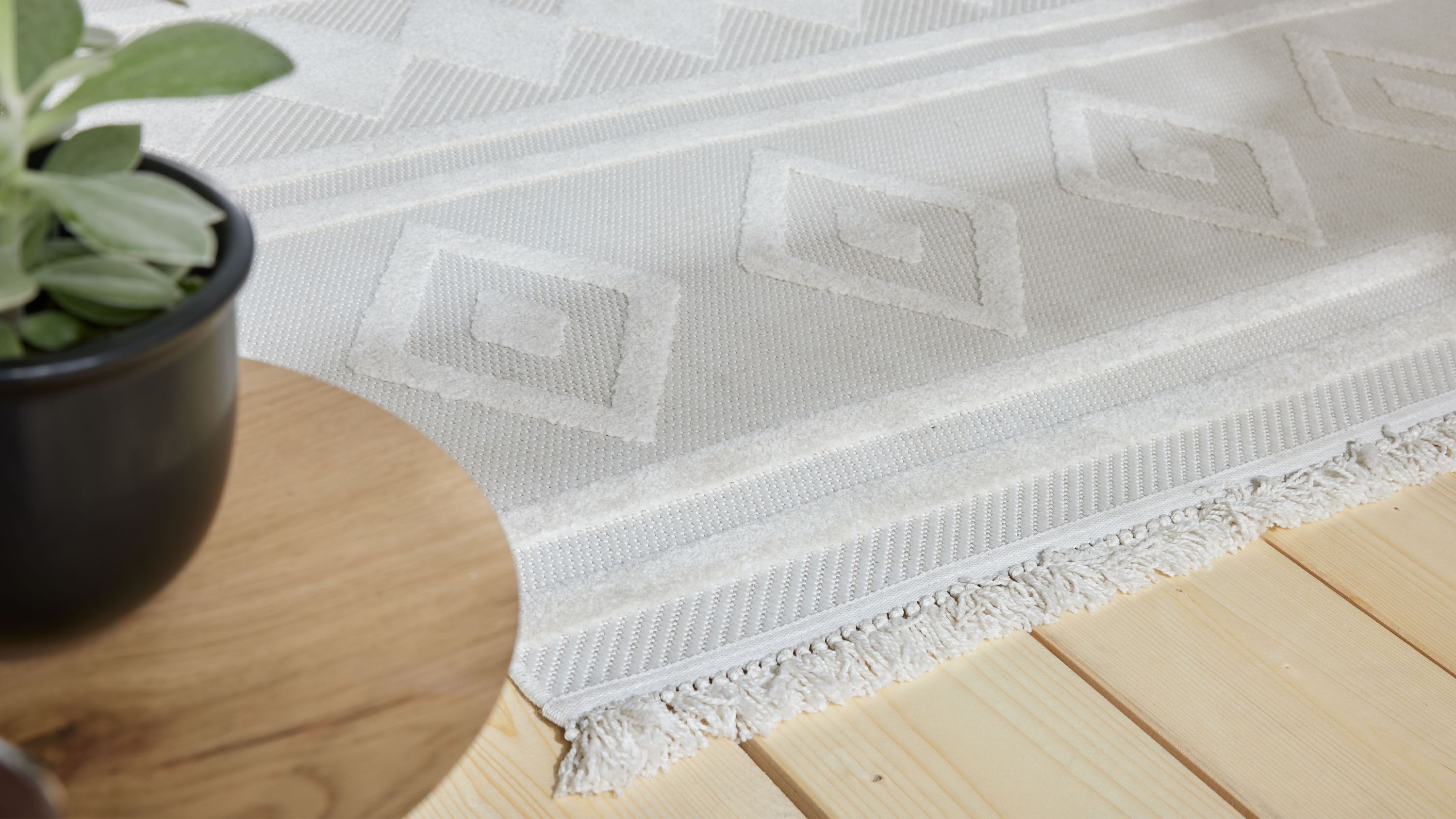 Beiger Teppich mit Rautenmuster und Fransen an der Kante auf hellem Holzboden neben einem schwarzen Blumentopf