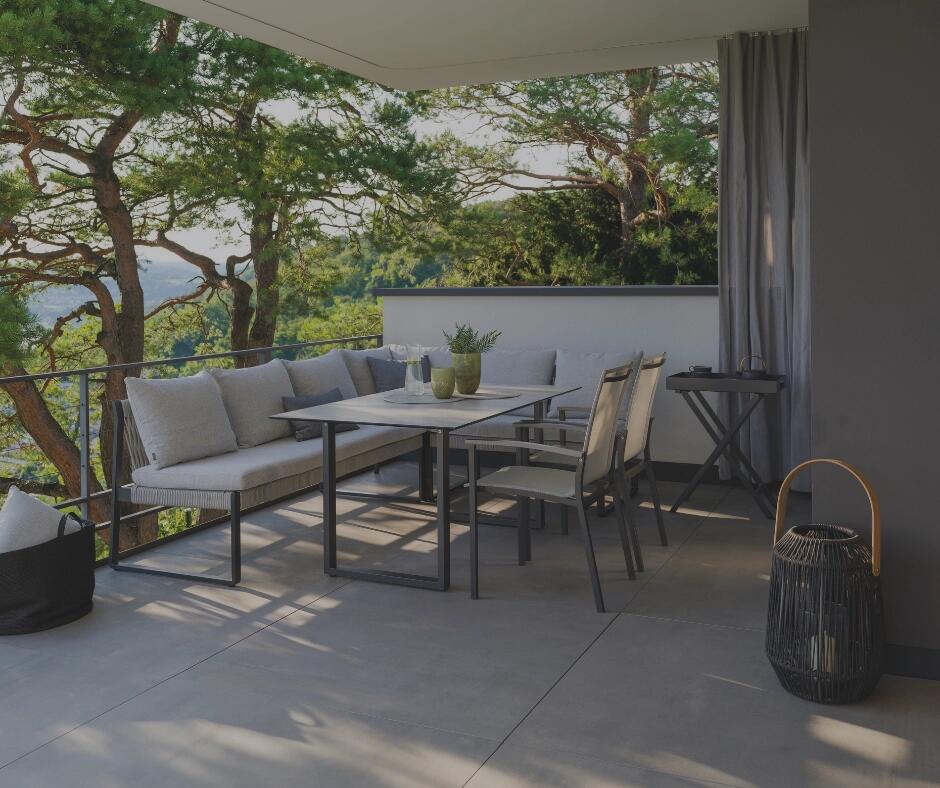 Überdachte Terrasse mit Outdoor-Eckbank, Esstisch und Gartenstühlen, umgeben von Bäumen mit Blick ins Grüne.